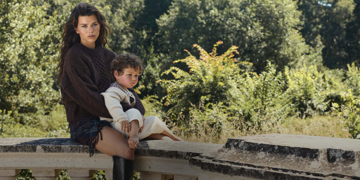A woman and a young child sit on a stone wall outdoors, with a lush green forest background. The woman wears a dark brown knit sweater and shorts, and the child is dressed in a cream-colored outfit.