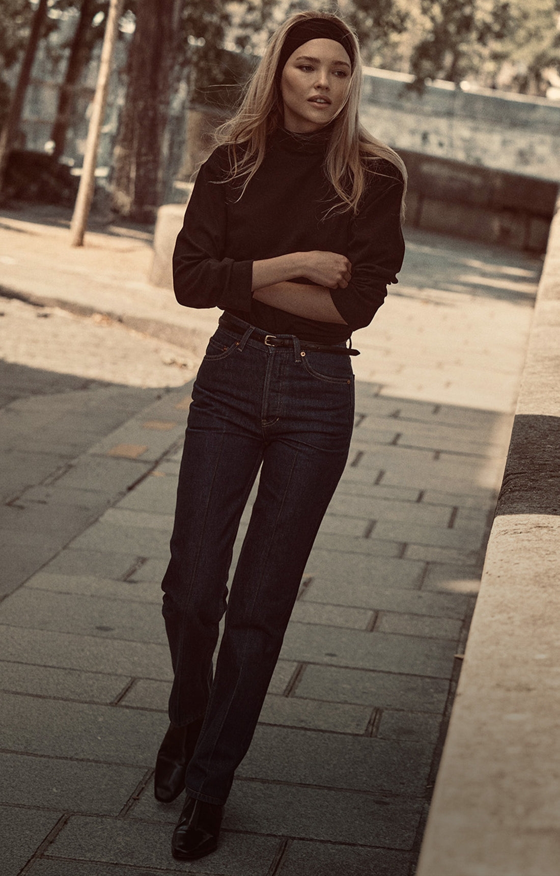 A woman wearing a black turtleneck, dark blue jeans, and black ankle boots stands on a paved outdoor walkway with her arms crossed.