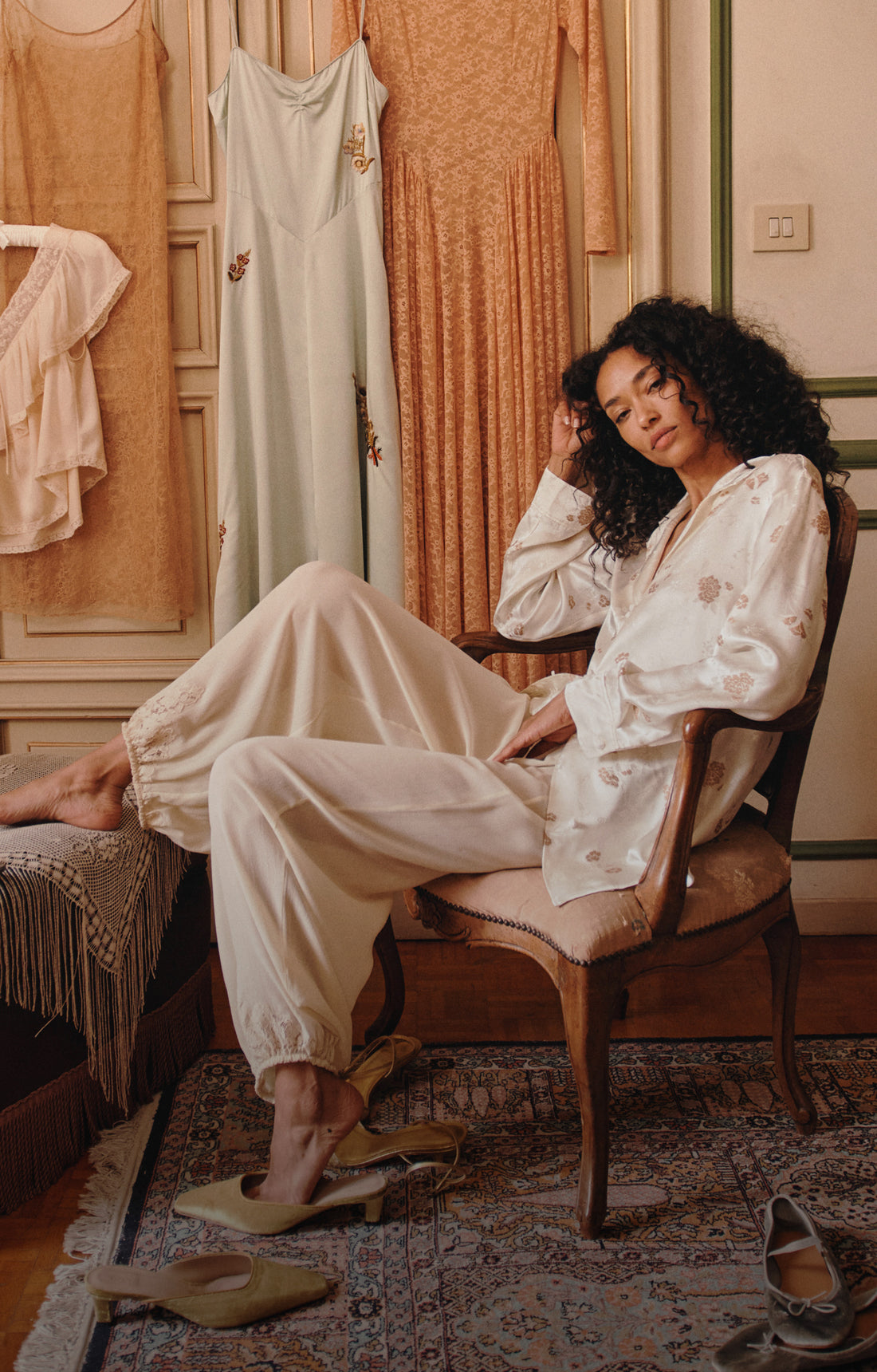 A woman with curly dark hair sits in a wooden armchair wearing a cream satin pajama set with embroidered details. The setting is a warm, vintage-style room with other pajamas hanging on a wardrobe, a patterned rug, and soft warm lighting.