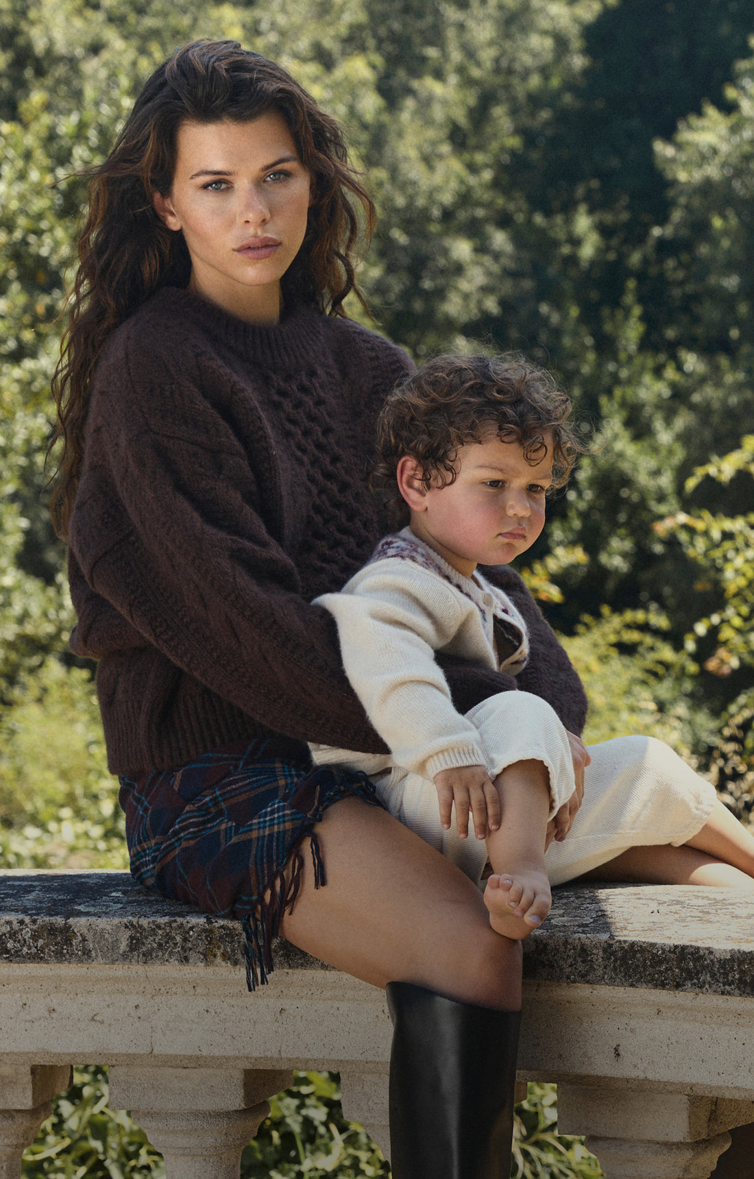 A woman and a young child sit together on a stone ledge outdoors, with trees in the background. The woman wears a dark brown cable-knit sweater, a dark plaid skirt, and tall black boots, while the child wears a cream knit sweater and cream pants.