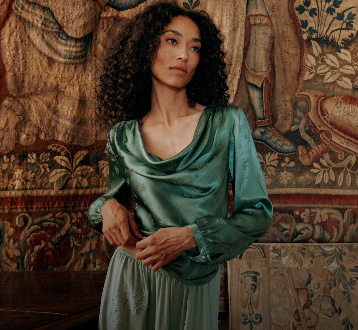 A woman with curly dark hair wearing a sage green satin cowl-neck top and matching skirt, standing in front of an ornate tapestry.