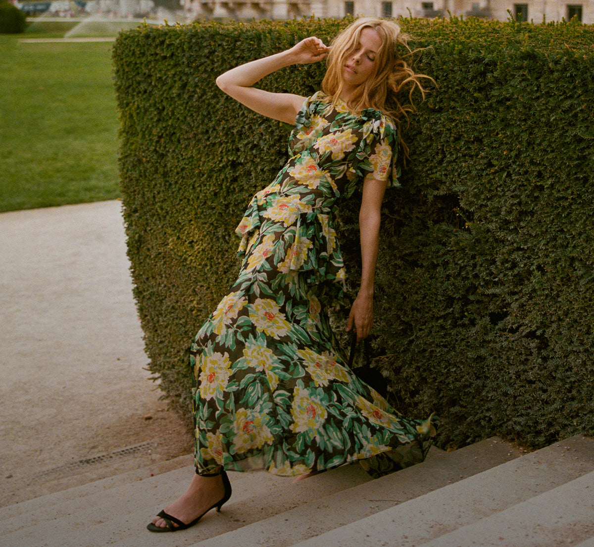 A woman wearing a green floral midi dress leans against a tall green hedge on outdoor stone steps, with grass and a building visible in the background.