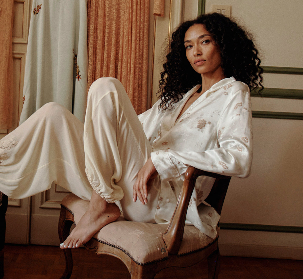 A woman with curly dark hair sits on a wooden chair in a warmly lit room, wearing a white satin pajama robe and matching pants with subtle floral embroidery.