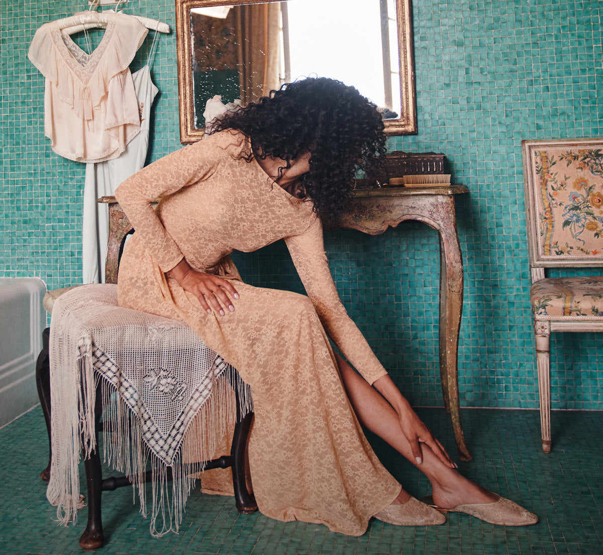 A woman with voluminous curly black hair sits on a chair in a vintage-style room with teal mosaic tile walls, wearing a beige lace long-sleeve maxi dress and beige slip-on shoes. A cream blouse hangs on a hanger on the left, and a wooden console table and floral chair are in the background.