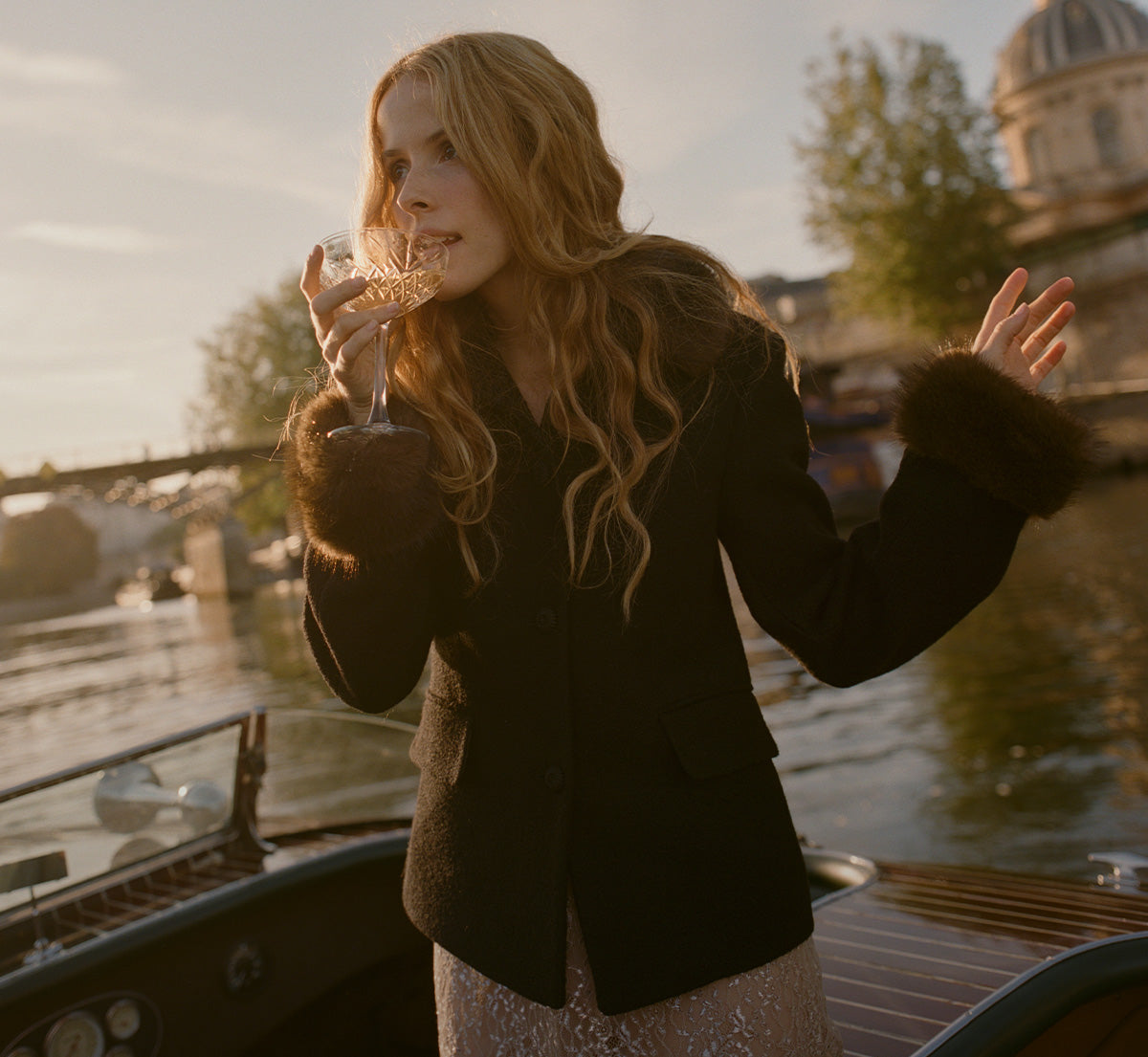 A woman with long wavy blonde hair stands on a boat by a canal at sunset, wearing a dark brown fur-trimmed coat and holding a glass of champagne.