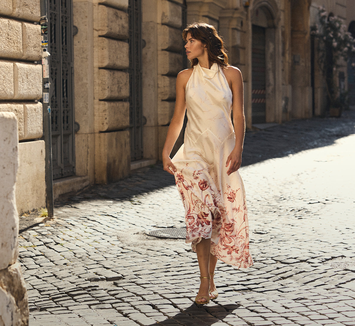 A woman walks down a sunlit cobblestone street wearing a sleeveless cream halter dress with a red and pink floral print on the lower skirt.