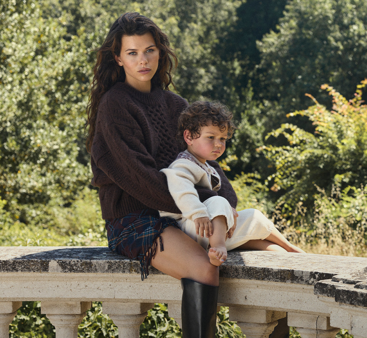 A woman and a young child sit on a stone bridge outdoors, with dense green foliage in the background. The woman wears a dark brown cable-knit sweater, a dark plaid skirt, and black knee-high boots, while holding the child who is dressed in a cream outfit.