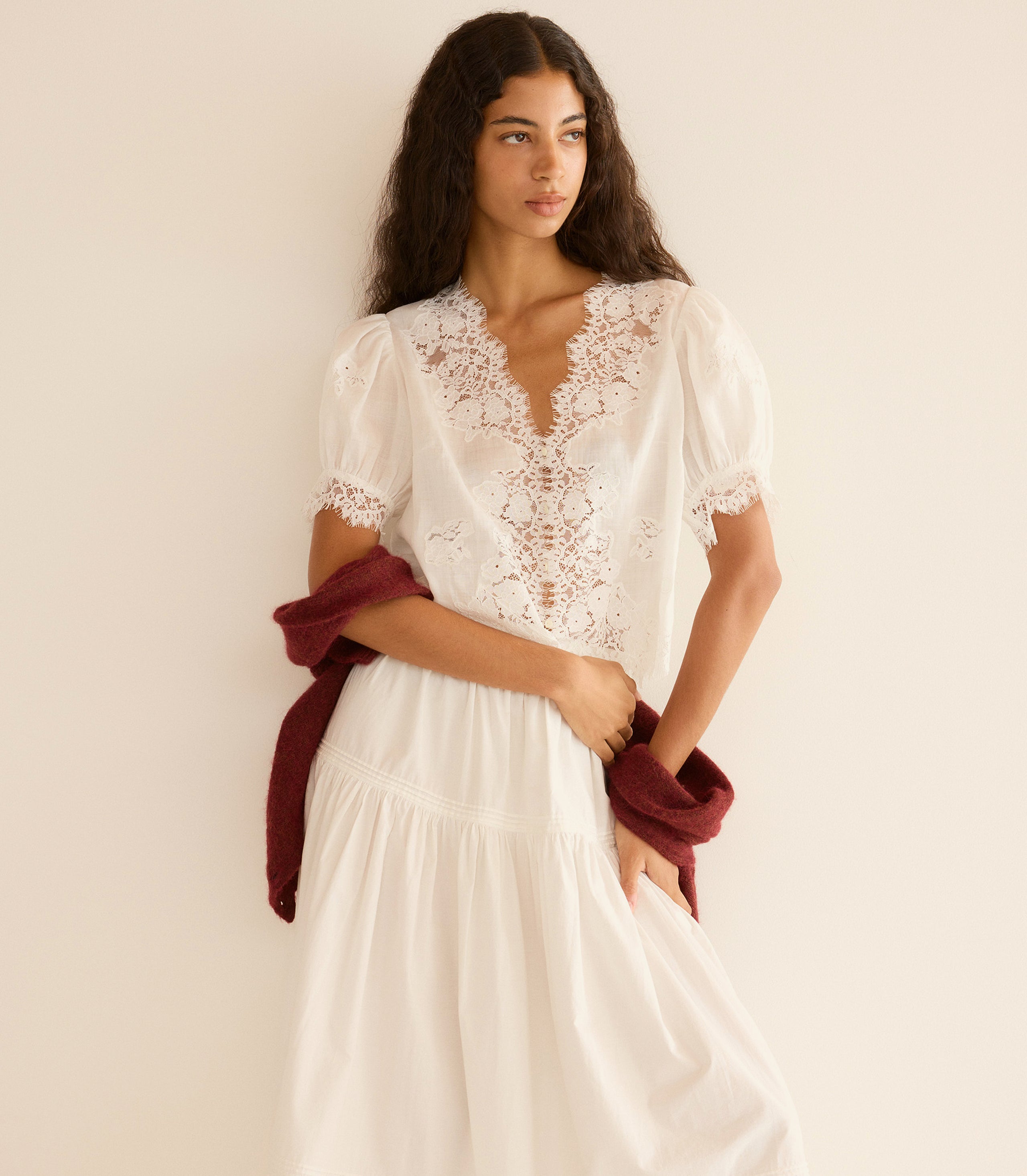 Woman wearing a white lace top and white skirt with a red shawl over her shoulders against a beige background