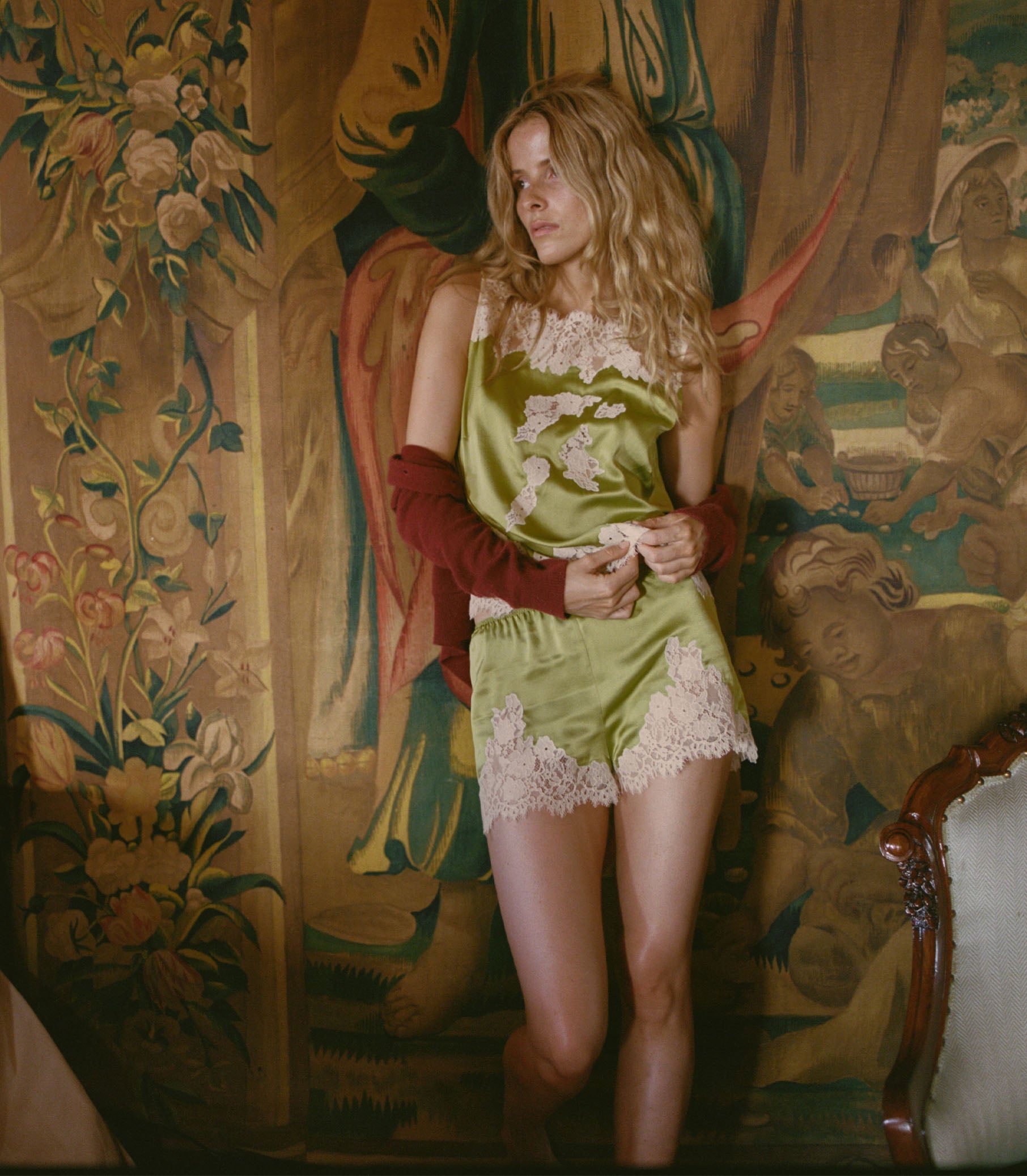 Woman in a green top and shorts with lace details standing in a room with ornate wallpaper.