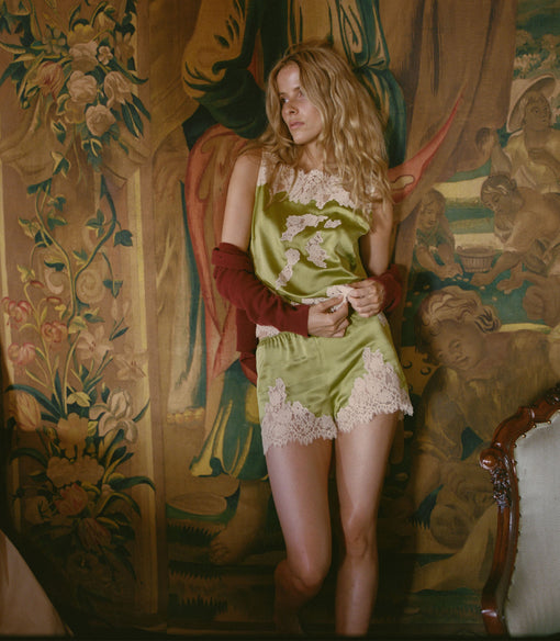 Woman in a green top and shorts with lace details standing in a room with ornate wallpaper.