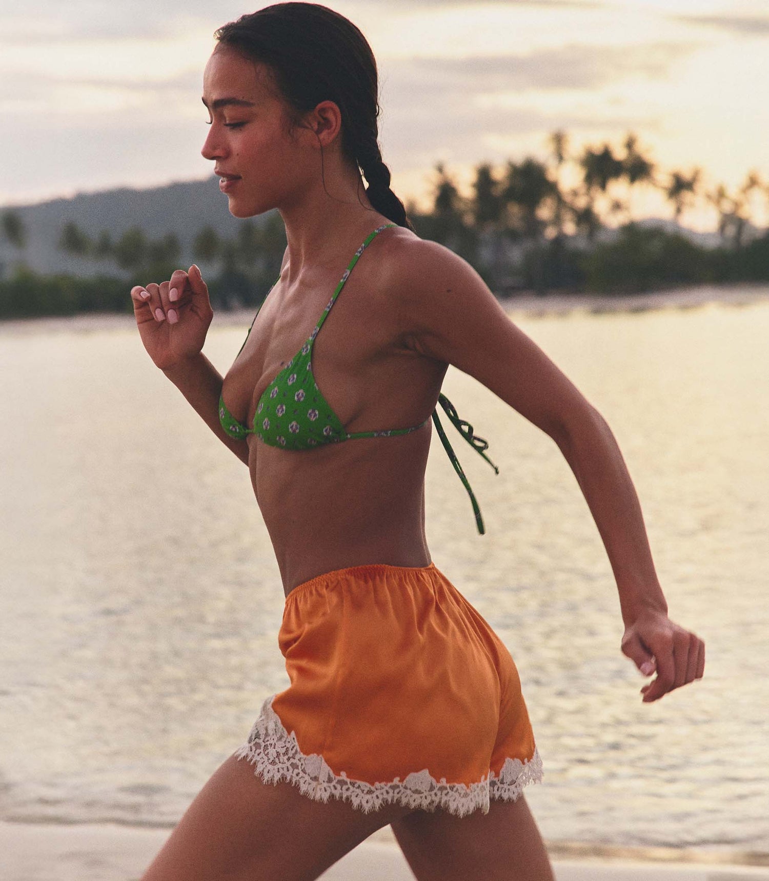 Woman in green bikini top and orange shorts standing on a beach at sunset.