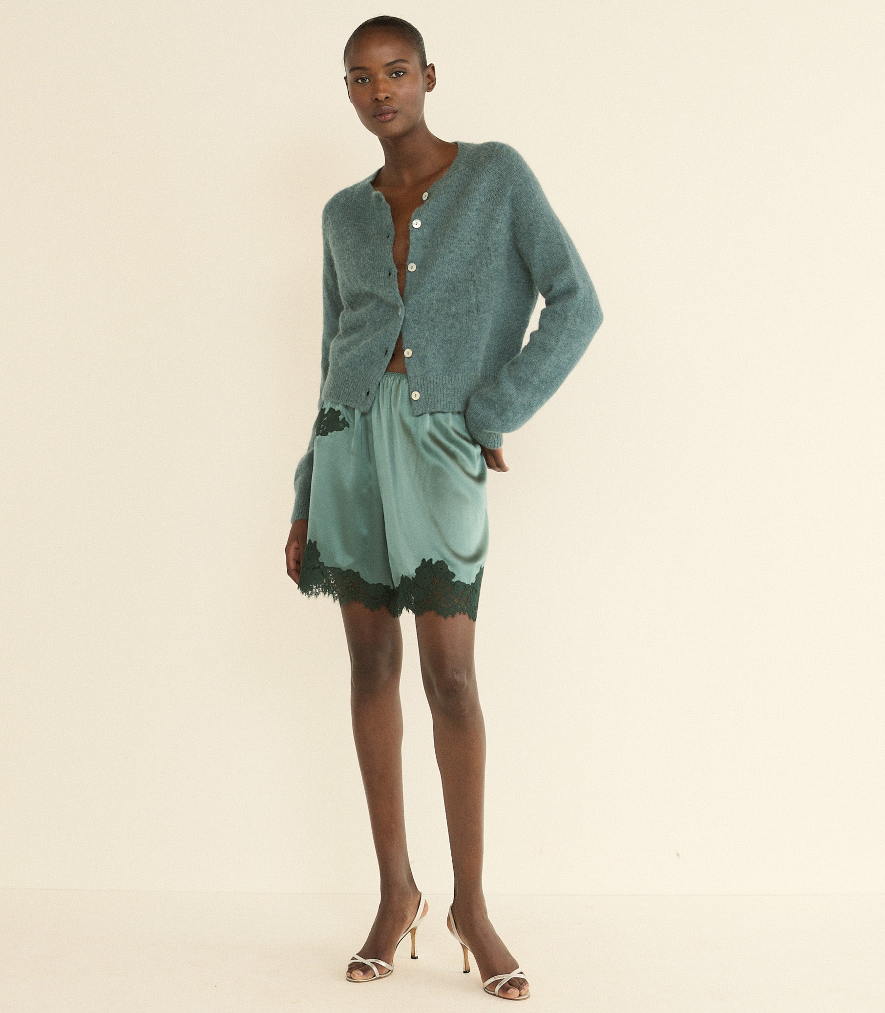 A model stands against a plain cream studio background wearing a sage green cardigan, a matching sage green satin pair of shorts with dark green lace trim, and beige high-heeled sandals.