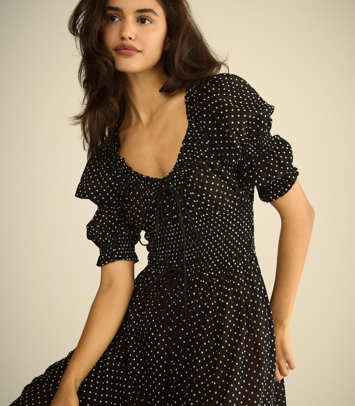 A woman wearing a black midi dress with white polka dots, featuring ruffled short sleeves, a deep scoop neckline, and a tie detail at the chest, posed against a solid beige background.
