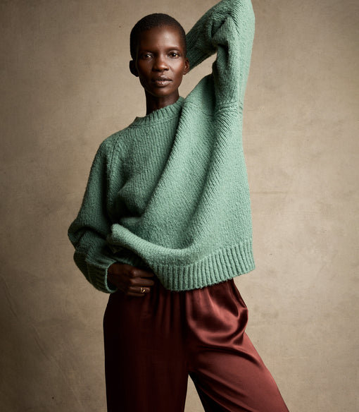 Person wearing a green sweater and maroon pants against a beige background