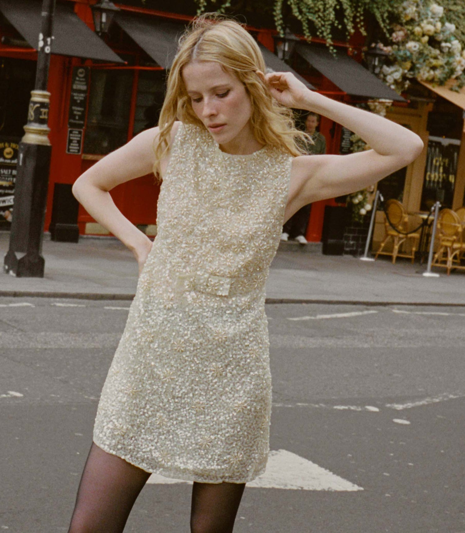 Woman in a gold sequin dress standing on a street with a building and chairs in the background.