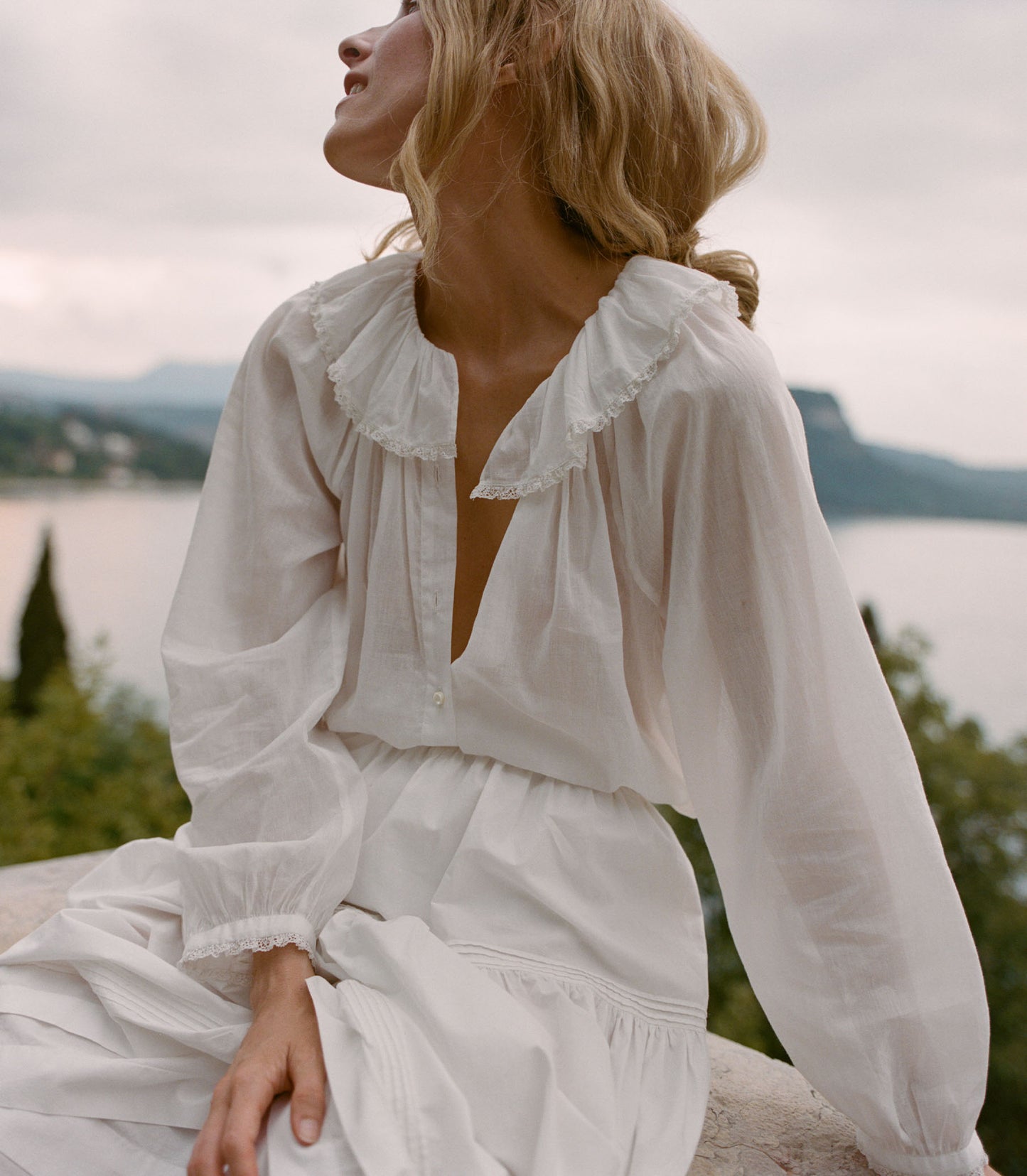 Woman in a white dress sitting by a lake with mountains in the background