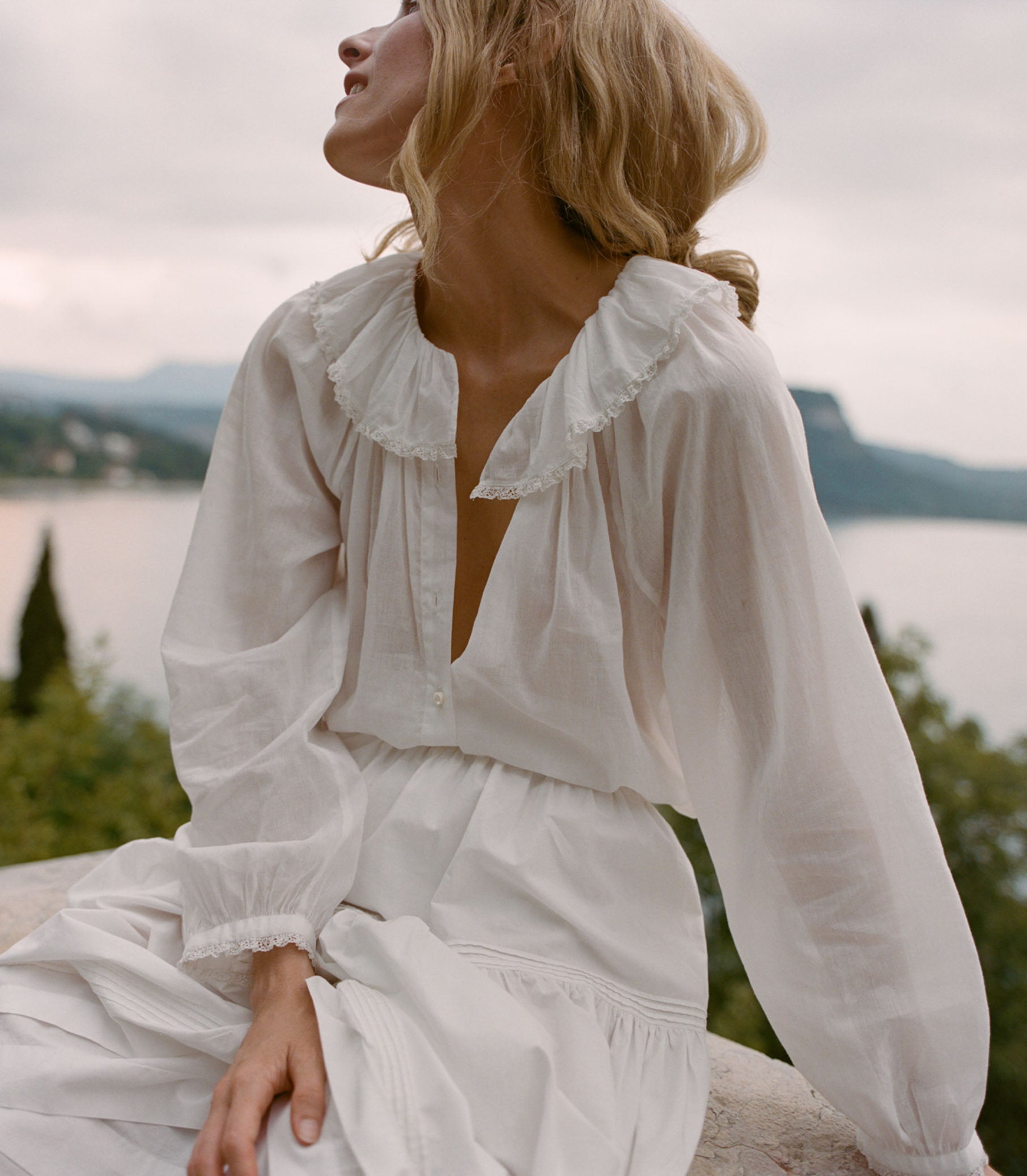 Woman in a white dress sitting by a lake with mountains in the background
