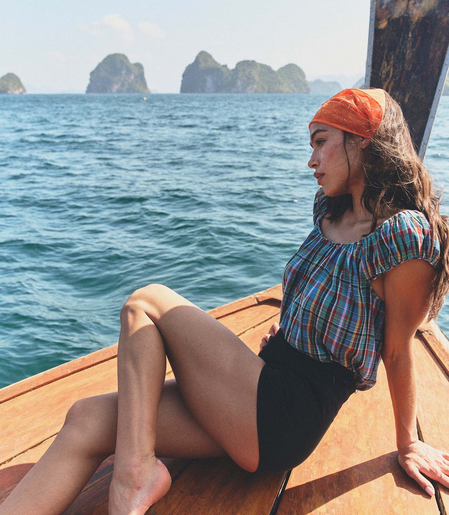 Woman wearing a plaid blouse, orange bandana, and black shorts sitting on a wooden boat with ocean and islands in the background
