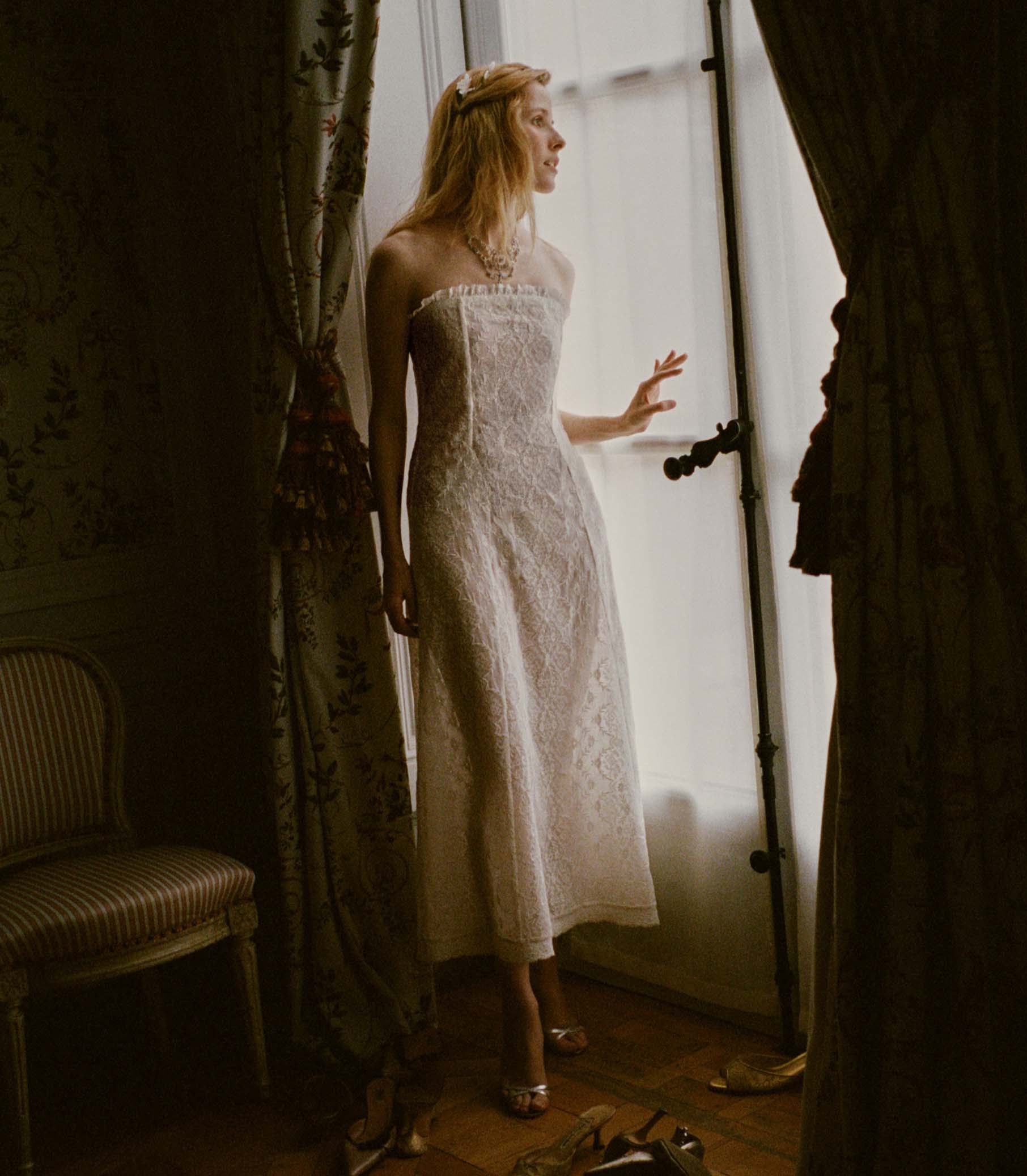 Woman in a strapless white lace dress standing in a room with curtains and a chair.
