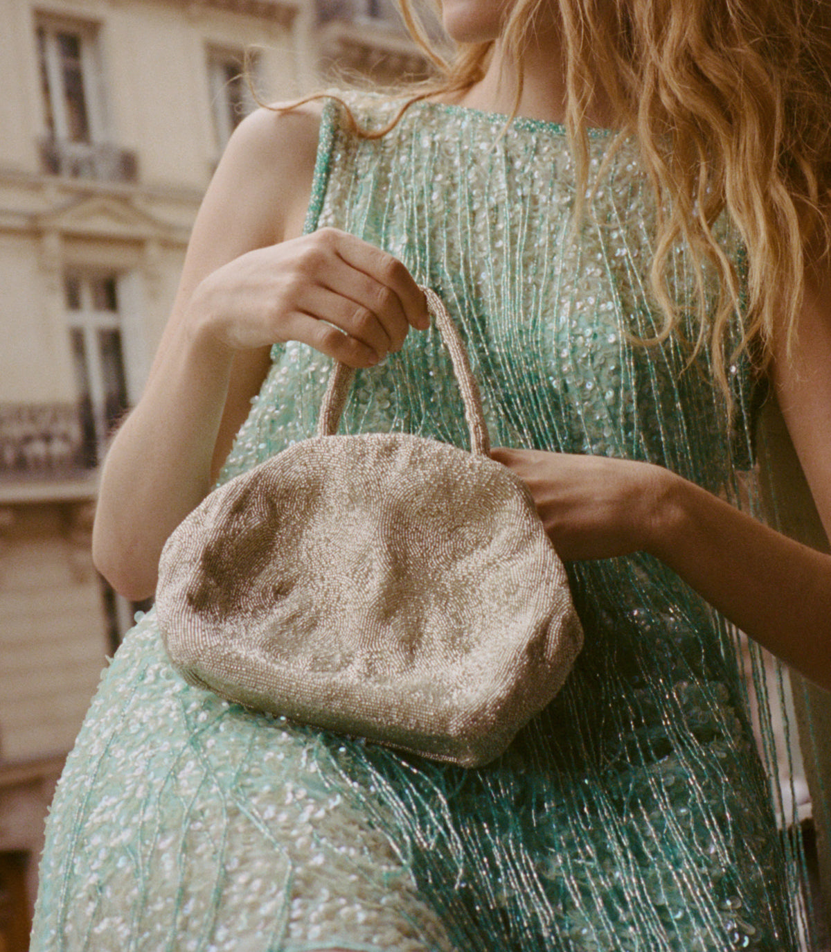 A woman in a shimmering mint green dress holds a small, textured metallic gold handbag with rope handles, set against a softly blurred urban background. 1
