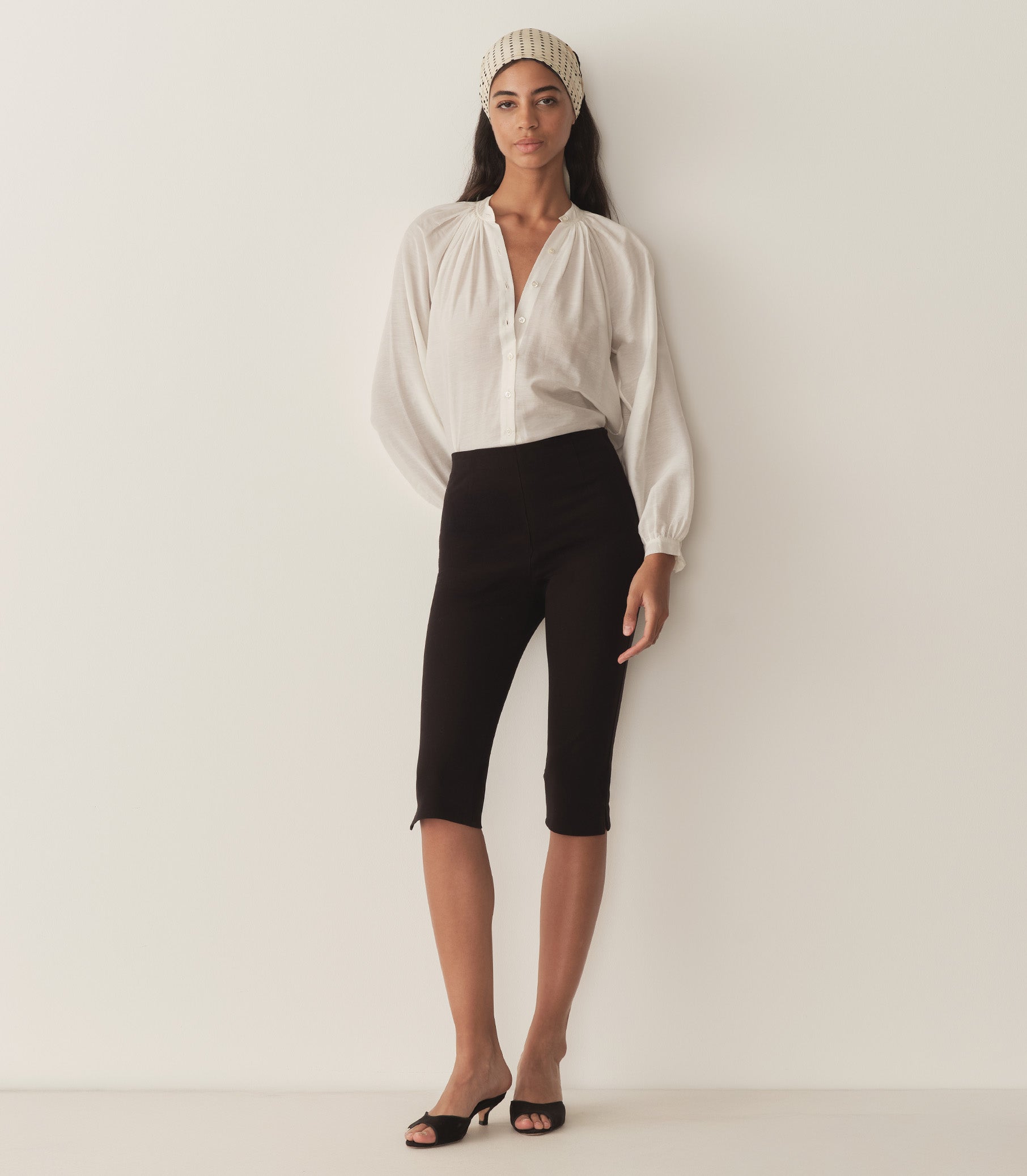 Rocio wears a white top with full-length raglan sleeves, a wide stand collar, and mother-of-pearl buttons at the center front and black capris with black heels.