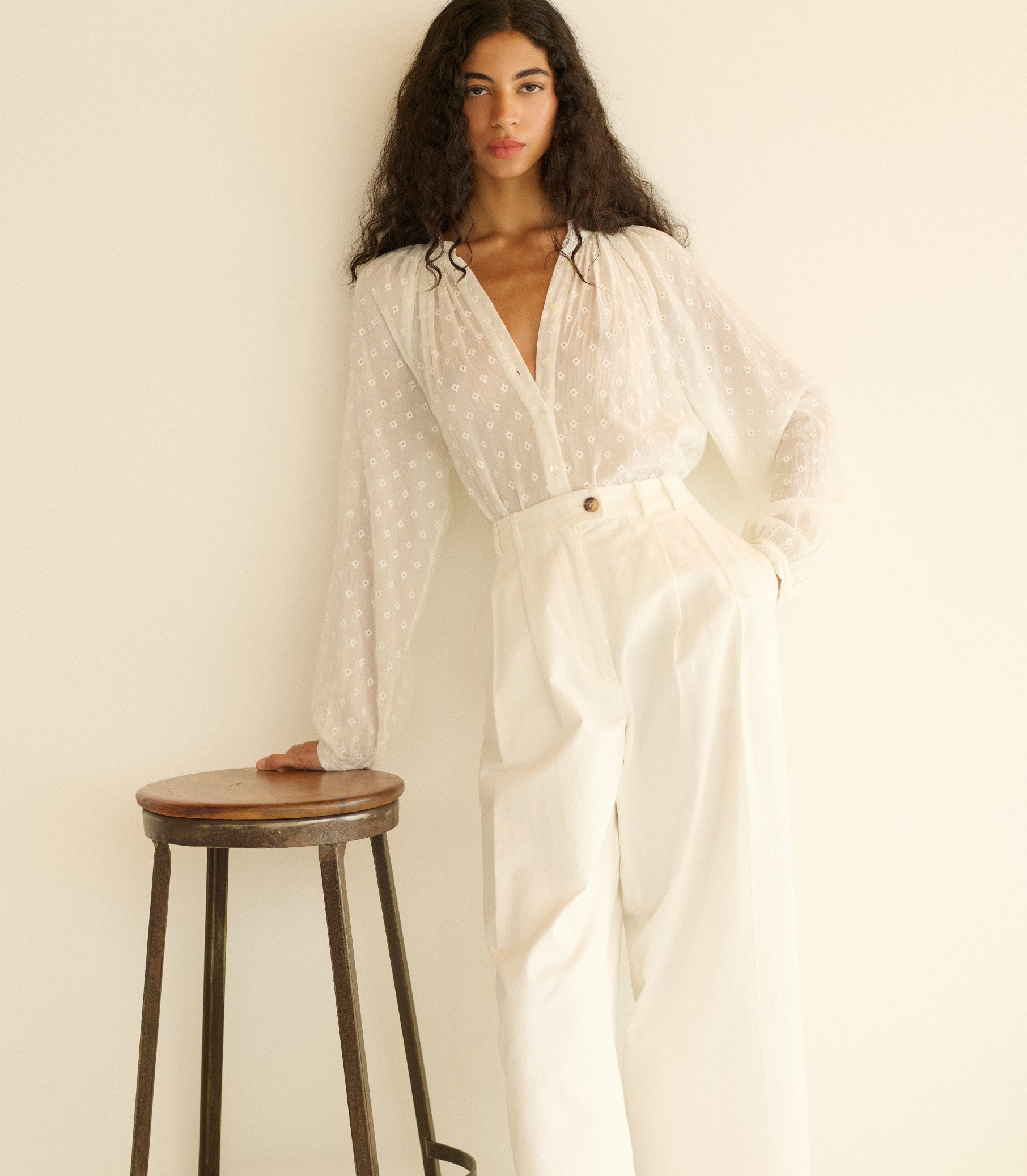 Woman wearing a white outfit standing next to a wooden stool against a plain background