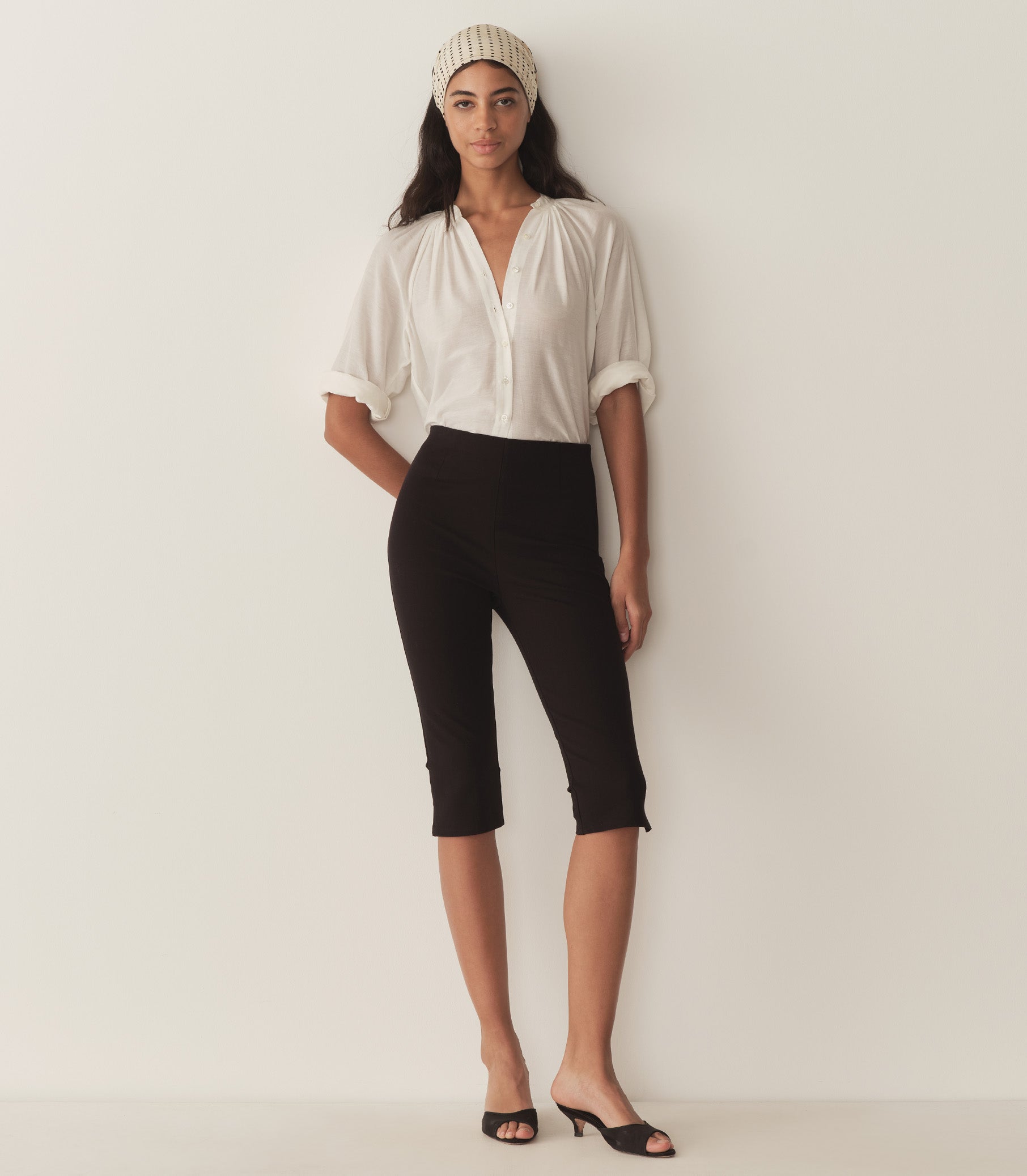 Rocio wears a white top with full-length raglan sleeves, a wide stand collar, and mother-of-pearl buttons at the center front and black capris with black heels.