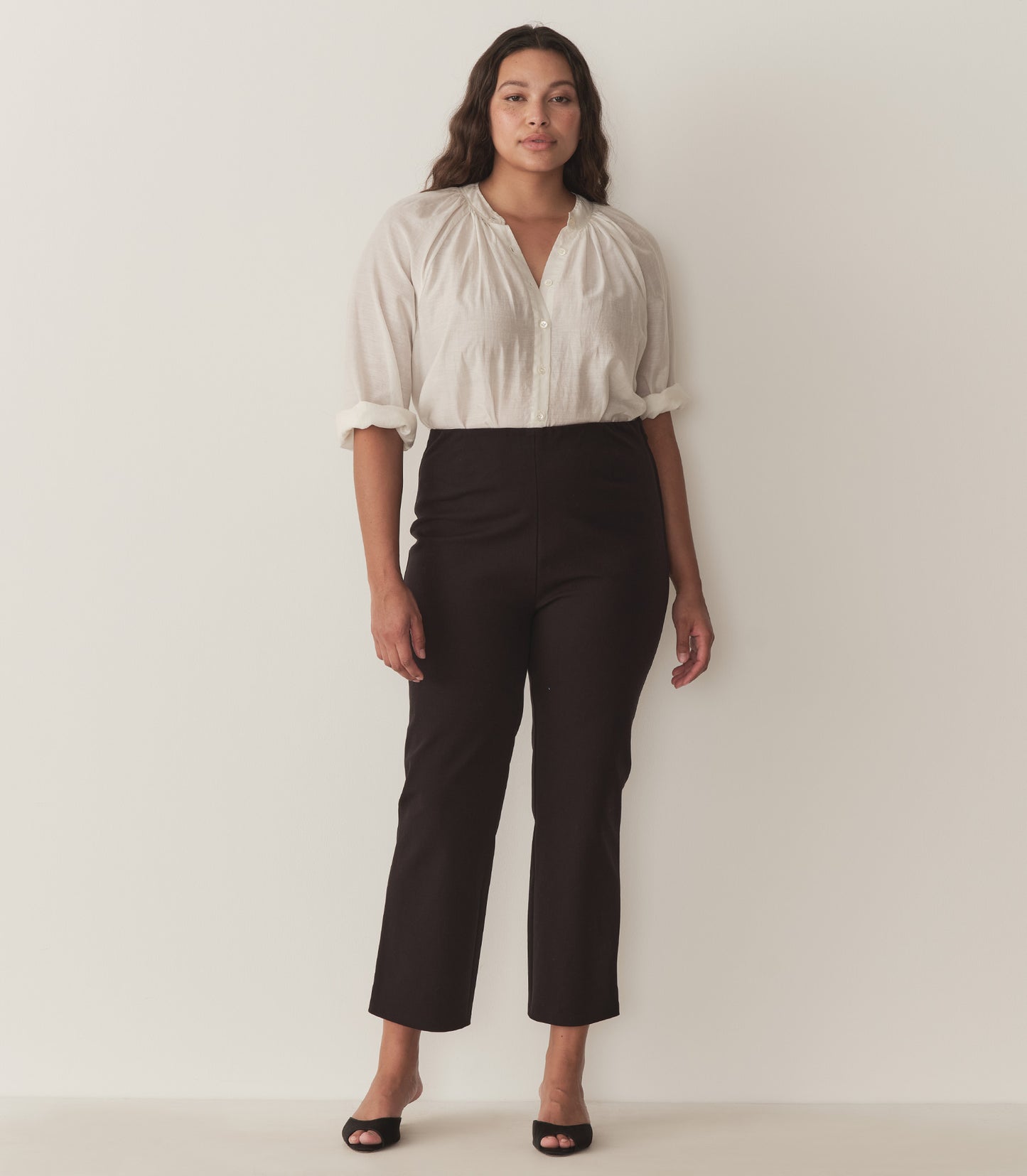 Alana wears a white top with full-length raglan sleeves, a wide stand collar, and mother-of-pearl buttons at the center front and black capris with black heels.