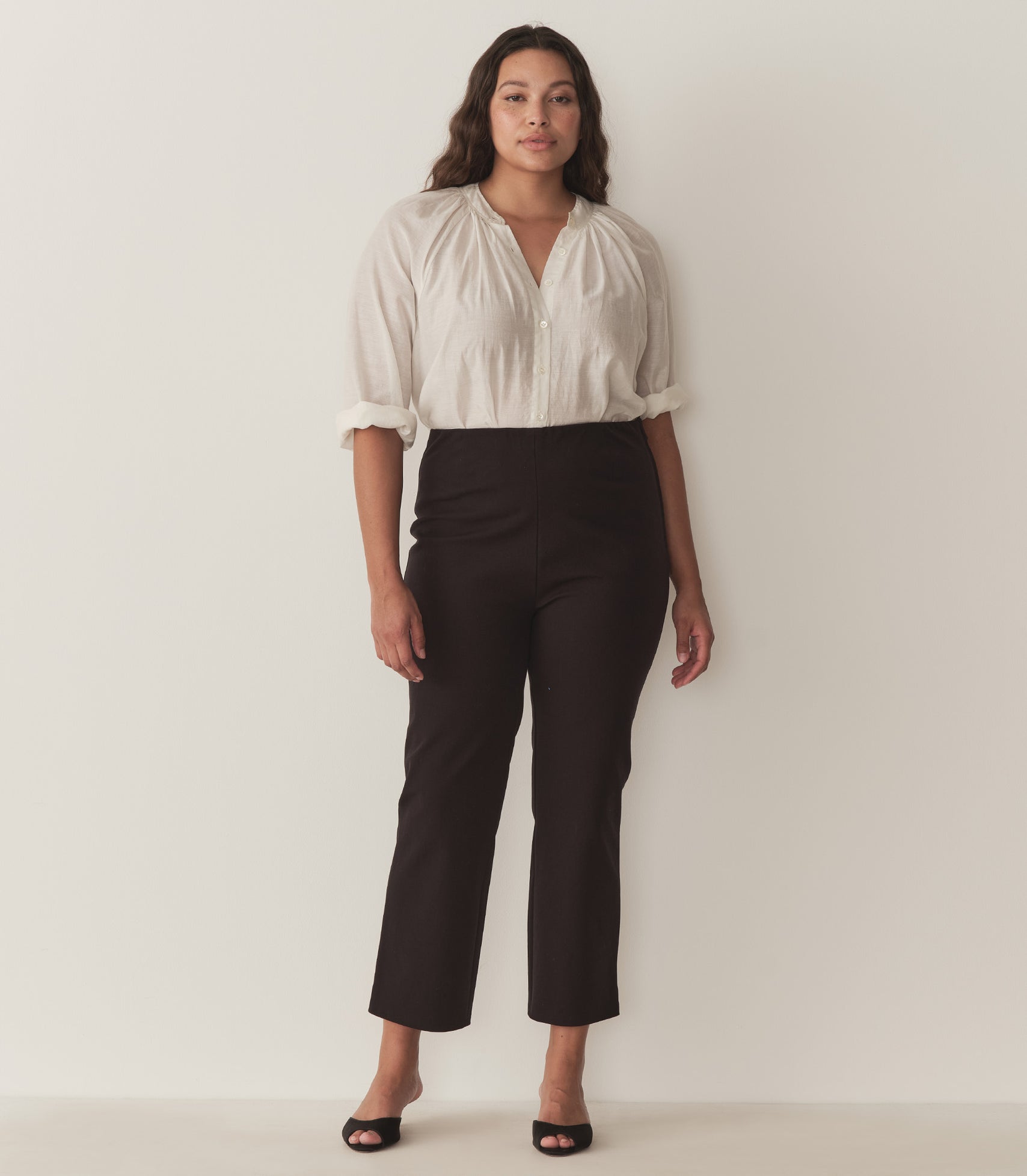 Alana wears a white top with full-length raglan sleeves, a wide stand collar, and mother-of-pearl buttons at the center front and black capris with black heels.