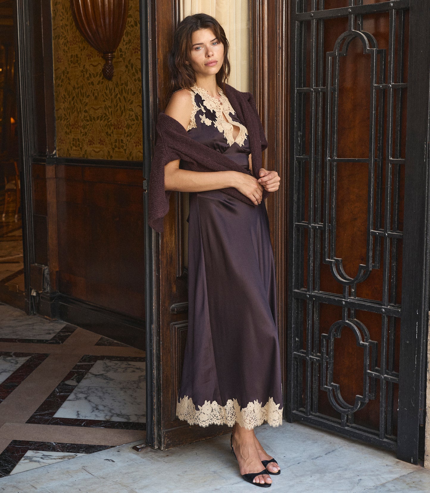A woman models a dark brown satin maxi dress with beige lace trim, styled with a matching draped shawl and a chunky beige necklace, standing in an elegant doorway.