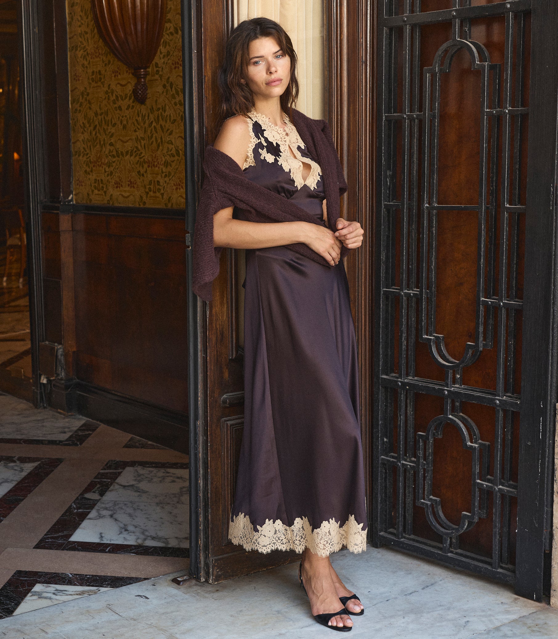 A woman models a dark brown satin maxi dress with beige lace trim, styled with a matching draped shawl and a chunky beige necklace, standing in an elegant doorway. 2
