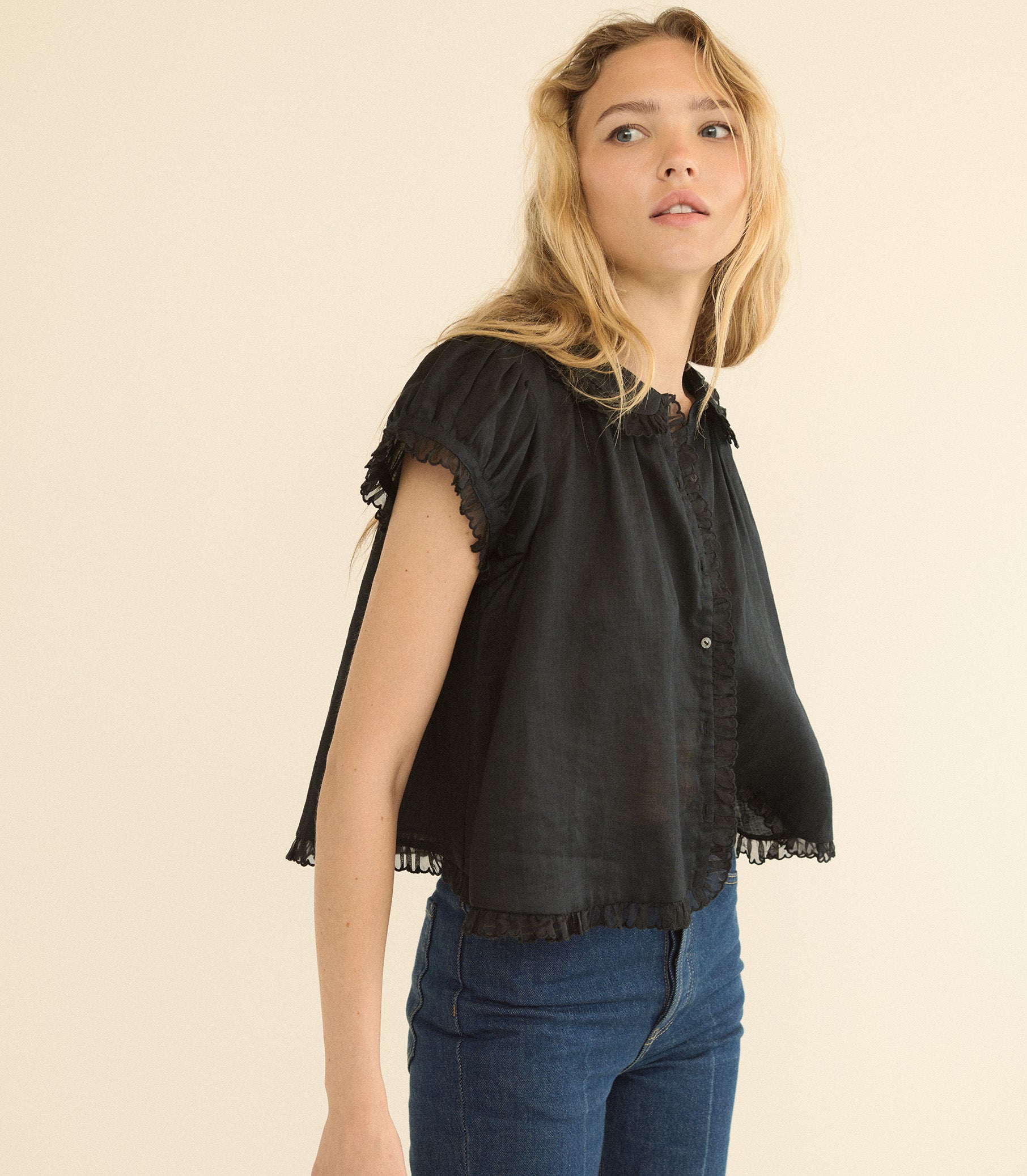 A woman wearing a black short-sleeve blouse with ruffled trim and blue jeans, photographed against a plain beige studio background. 1
