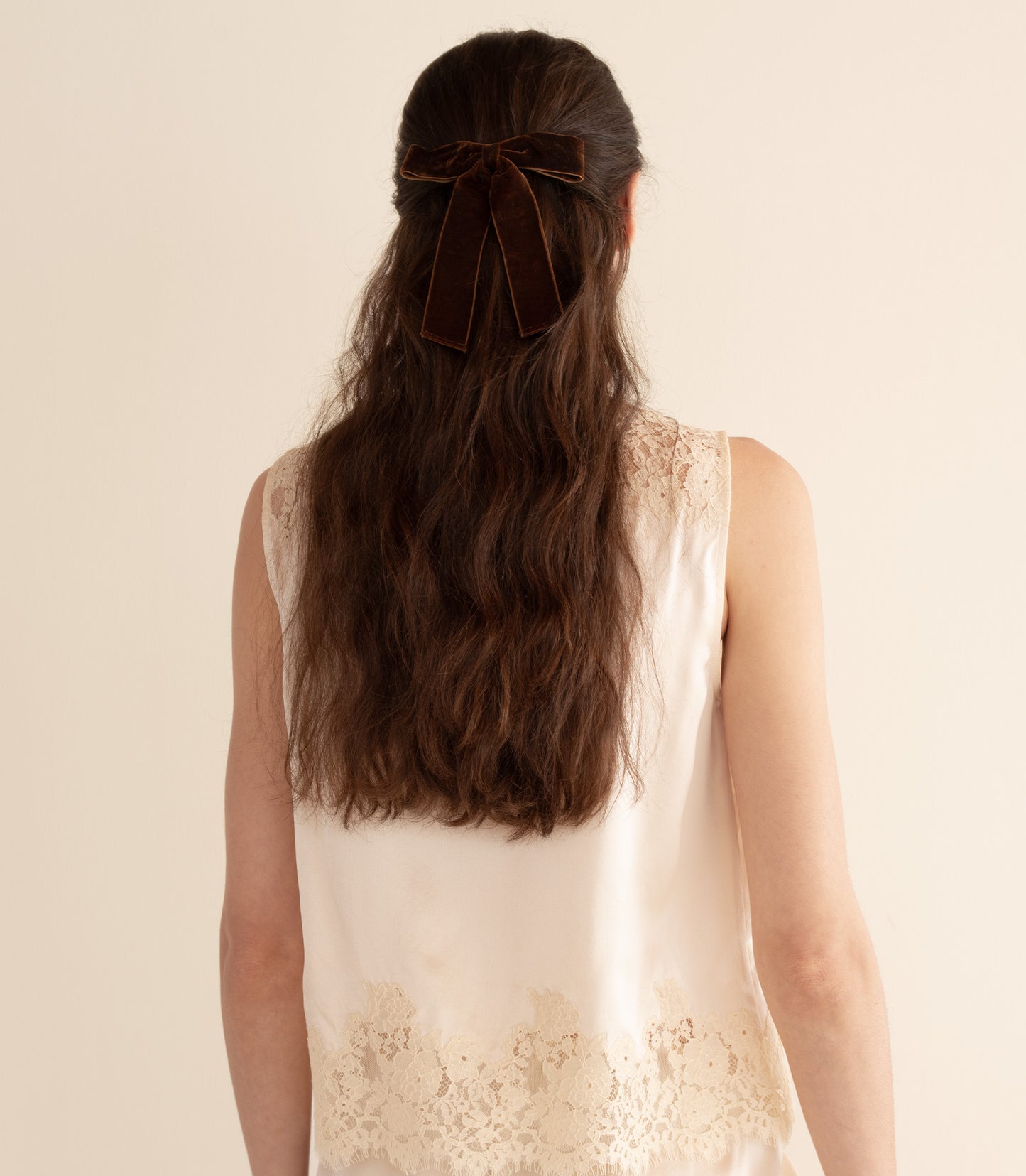 Woman with long hair styled with a bow, wearing a white lace top on a beige background