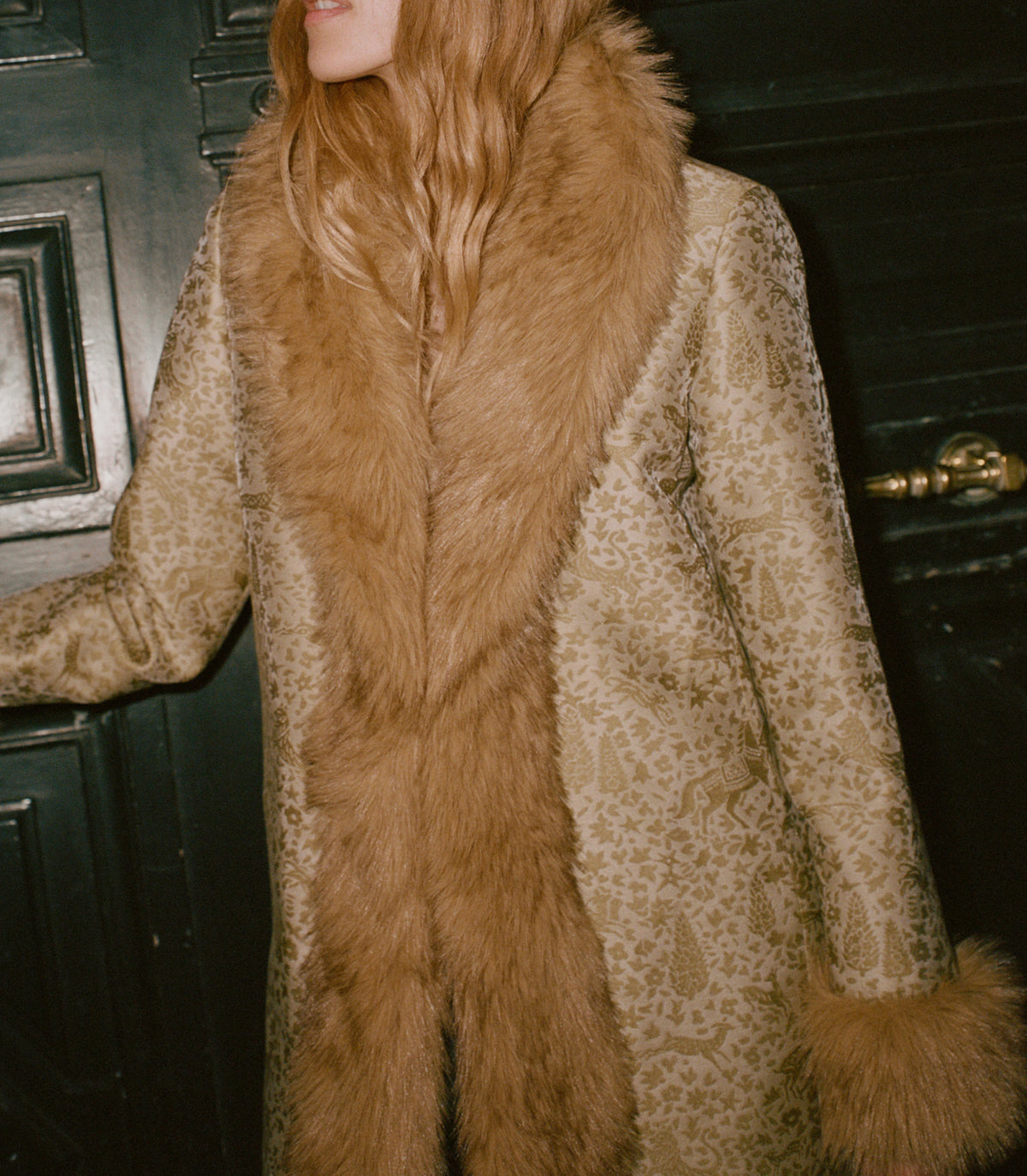 A person wearing a long beige and brown patterned coat with a large warm brown faux-fur collar and matching fur trim, standing in front of a dark wooden door. 1
