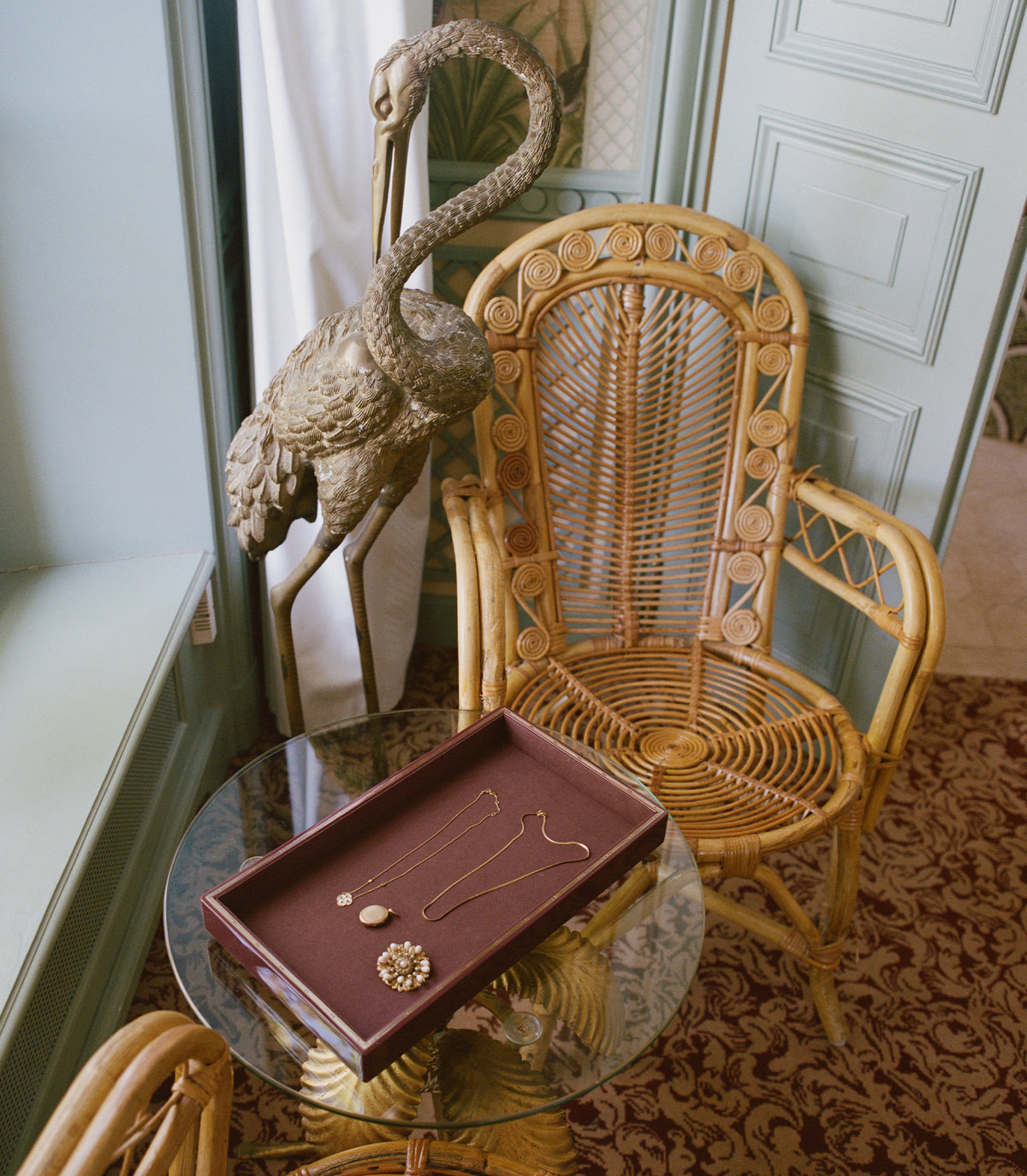 A styled interior scene with a rattan armchair, a glass-top side table holding a dark brown jewelry tray with gold necklaces and earrings, a decorative bird statue, and a pale paneled door in the background.