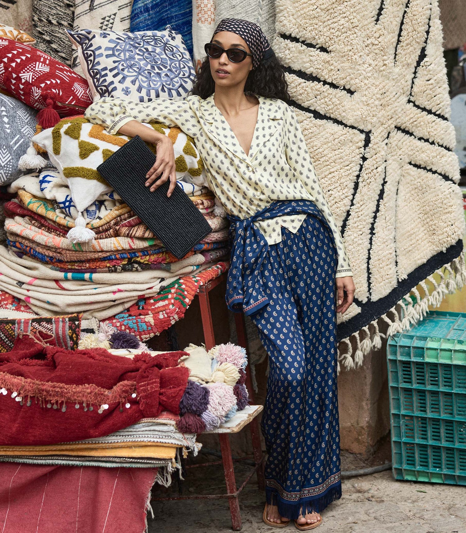 Woman in a paisley blouse and pants in a  market setting surrounded by colorful fabrics and rugs