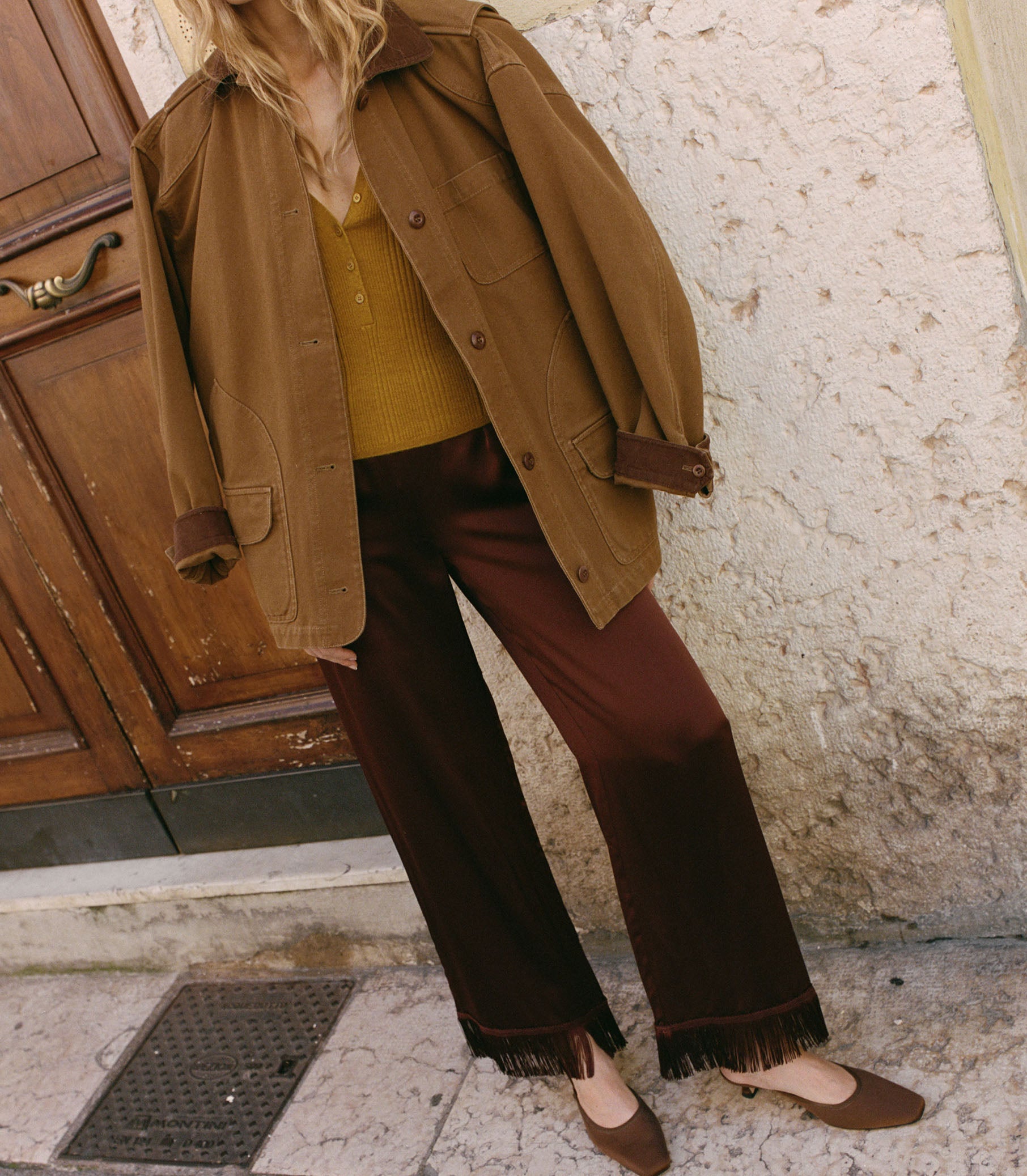 Person wearing a brown jacket, yellow top, and brown pants standing against a textured wall.
