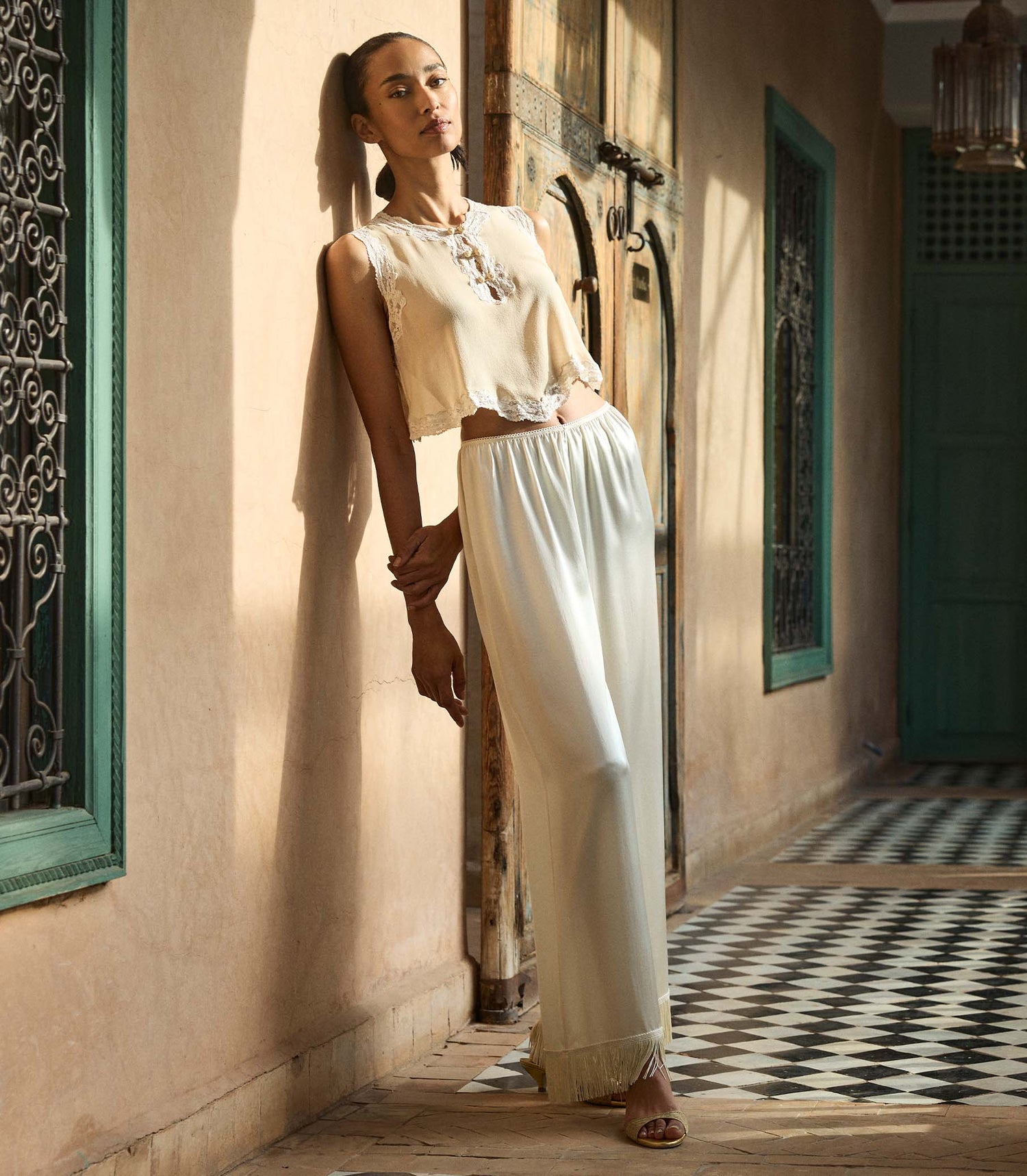 Woman in a blouse and white fringe pants standing in a sunlit hallway with decorative elements.