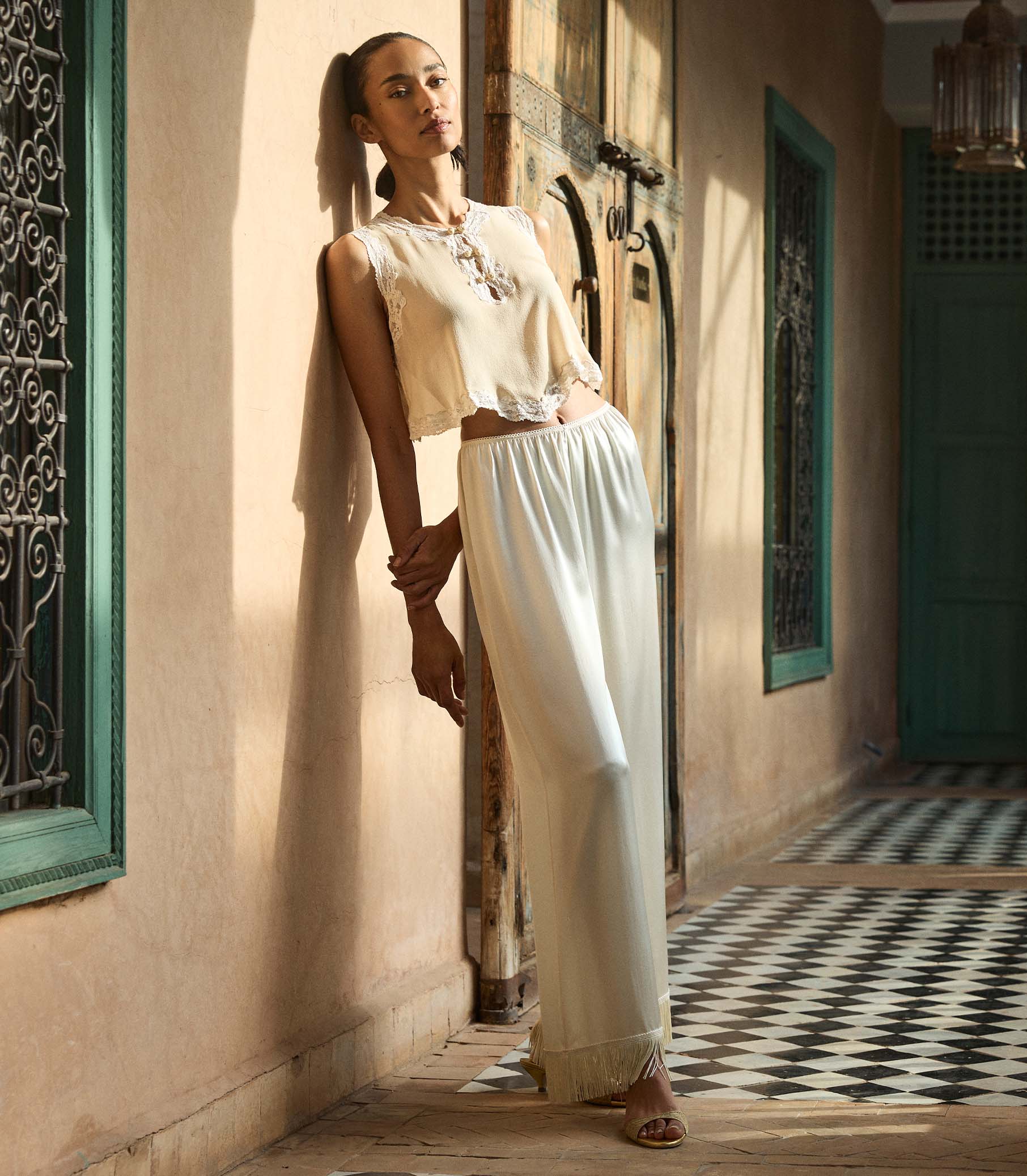 Woman in a blouse and white fringe pants standing in a sunlit hallway with decorative elements. 1
