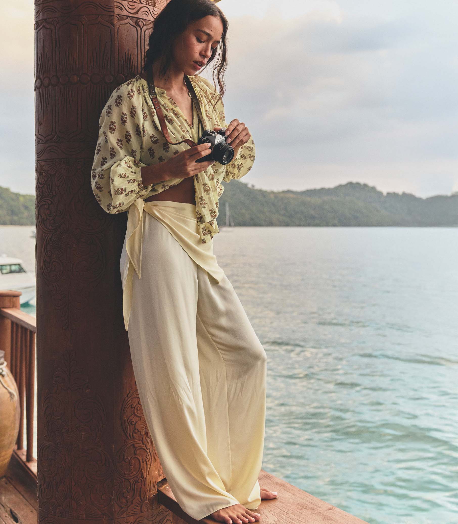 Woman in a yellow top and white pants holding a camera by a waterfront.
