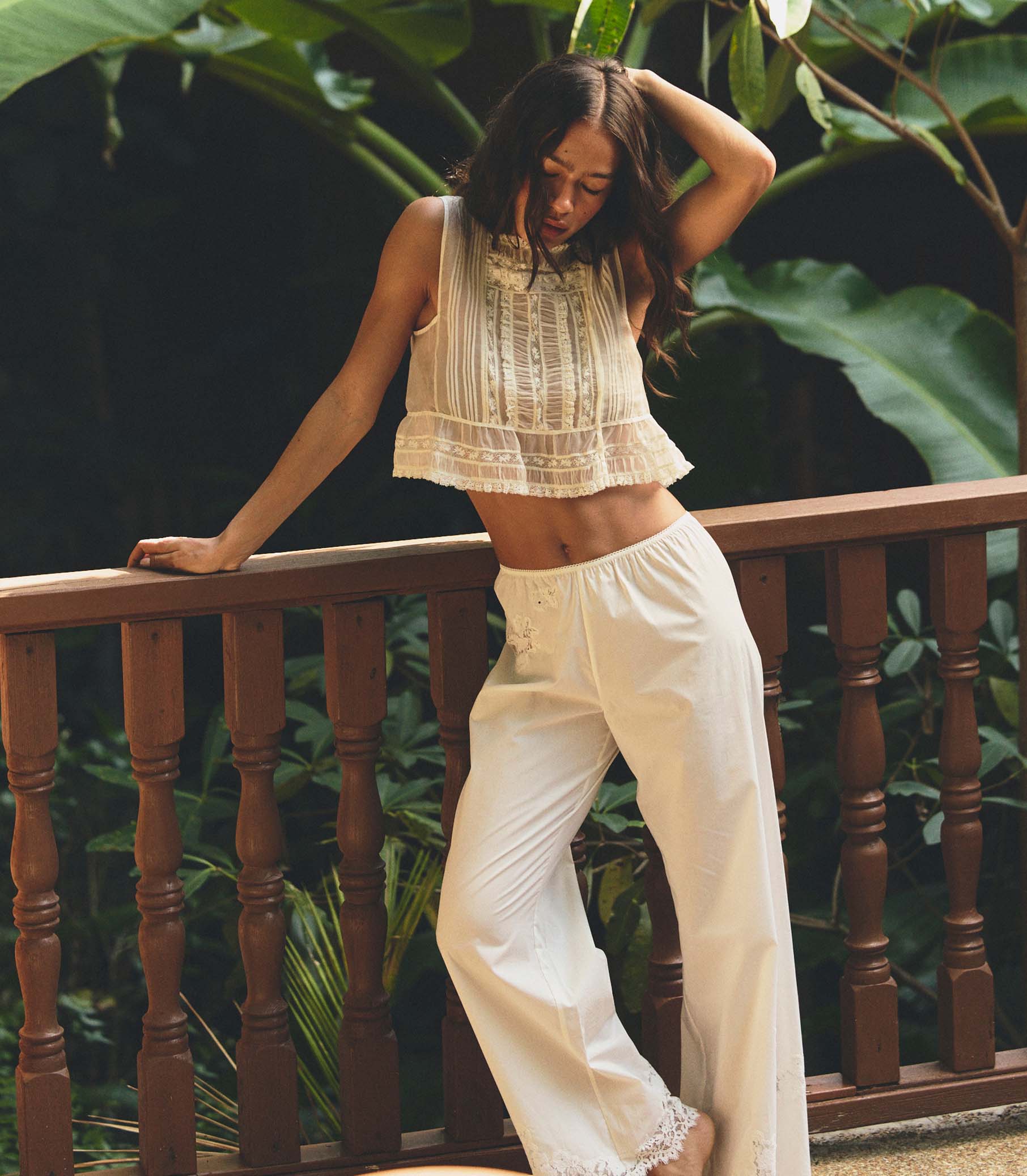 Woman in a white lace top and white pants standing on a wooden balcony with green foliage in the background