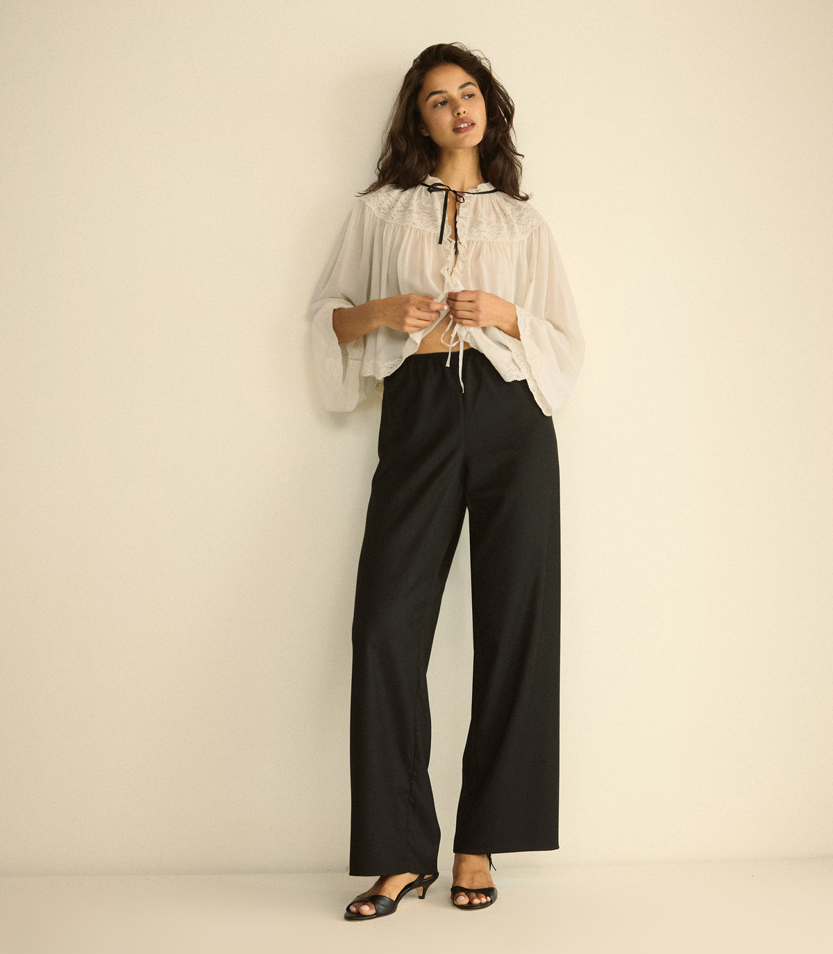 A woman stands against a plain cream studio wall wearing a sheer white long-sleeve blouse with a tie neckline and black wide-leg trousers.
