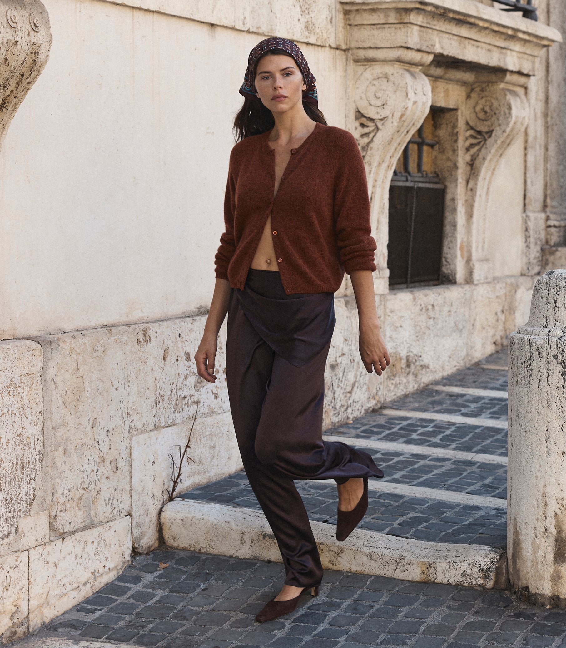 A woman wearing a rust-brown cardigan, dark brown draped trousers, and brown flats walks down a stone-paved street next to a beige stucco building.