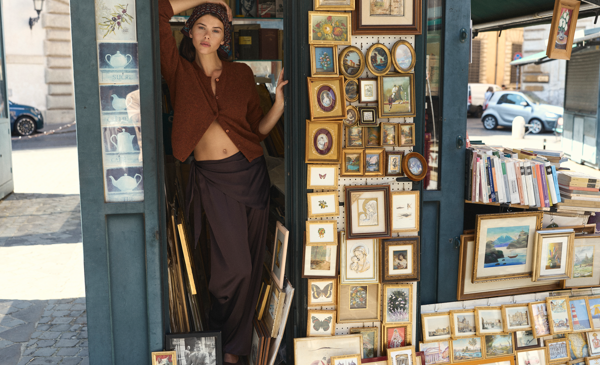 A woman in a printed headscarf leans in the doorway of a street kiosk filled with framed prints, wearing a cropped rust-brown cardigan and dark brown draped wide-leg trousers.