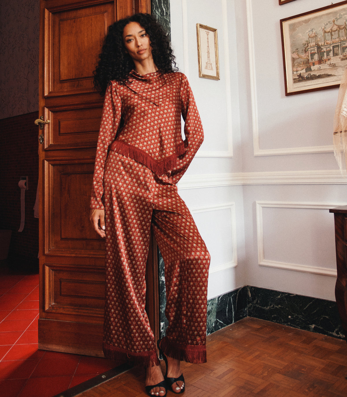 A woman models a rust-orange and white patterned two-piece set consisting of a long-sleeve top and matching wide-leg pants with fringe details, standing in an elegant room with wooden paneling and a dark wood door.