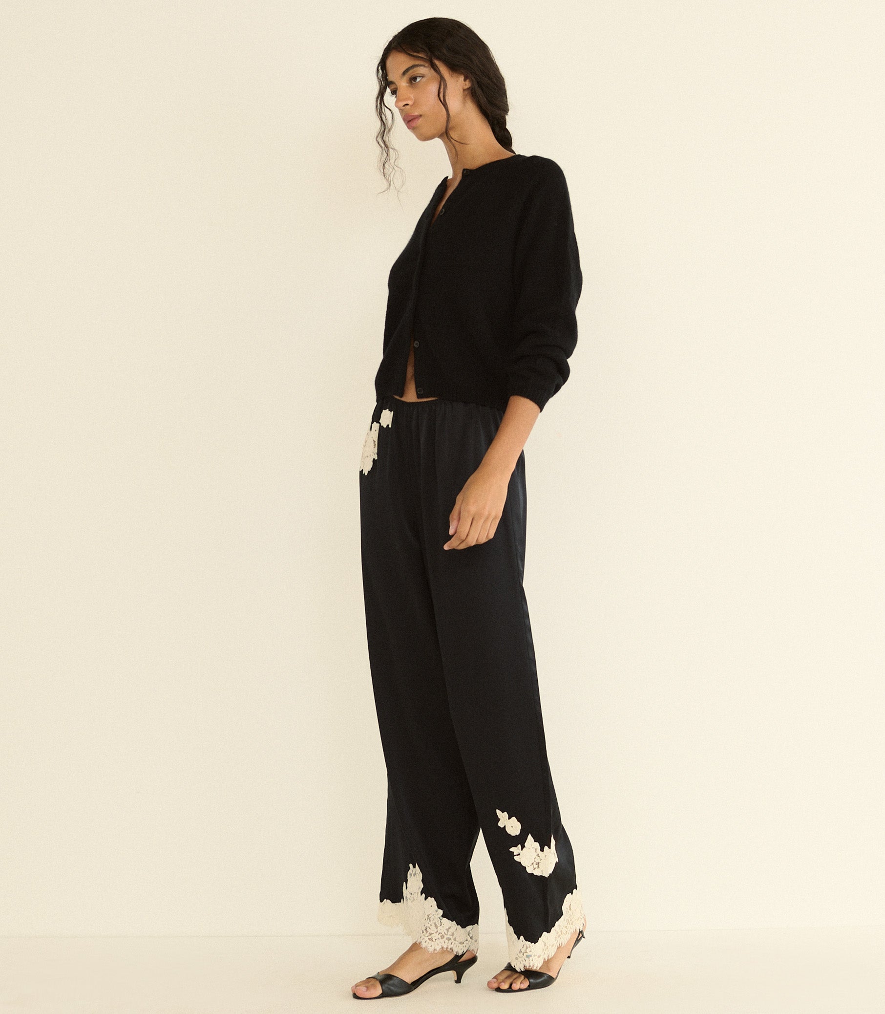 A woman models a black cardigan and wide-leg black trousers with white floral lace trim at the hems, paired with black heeled sandals, against a plain cream background.