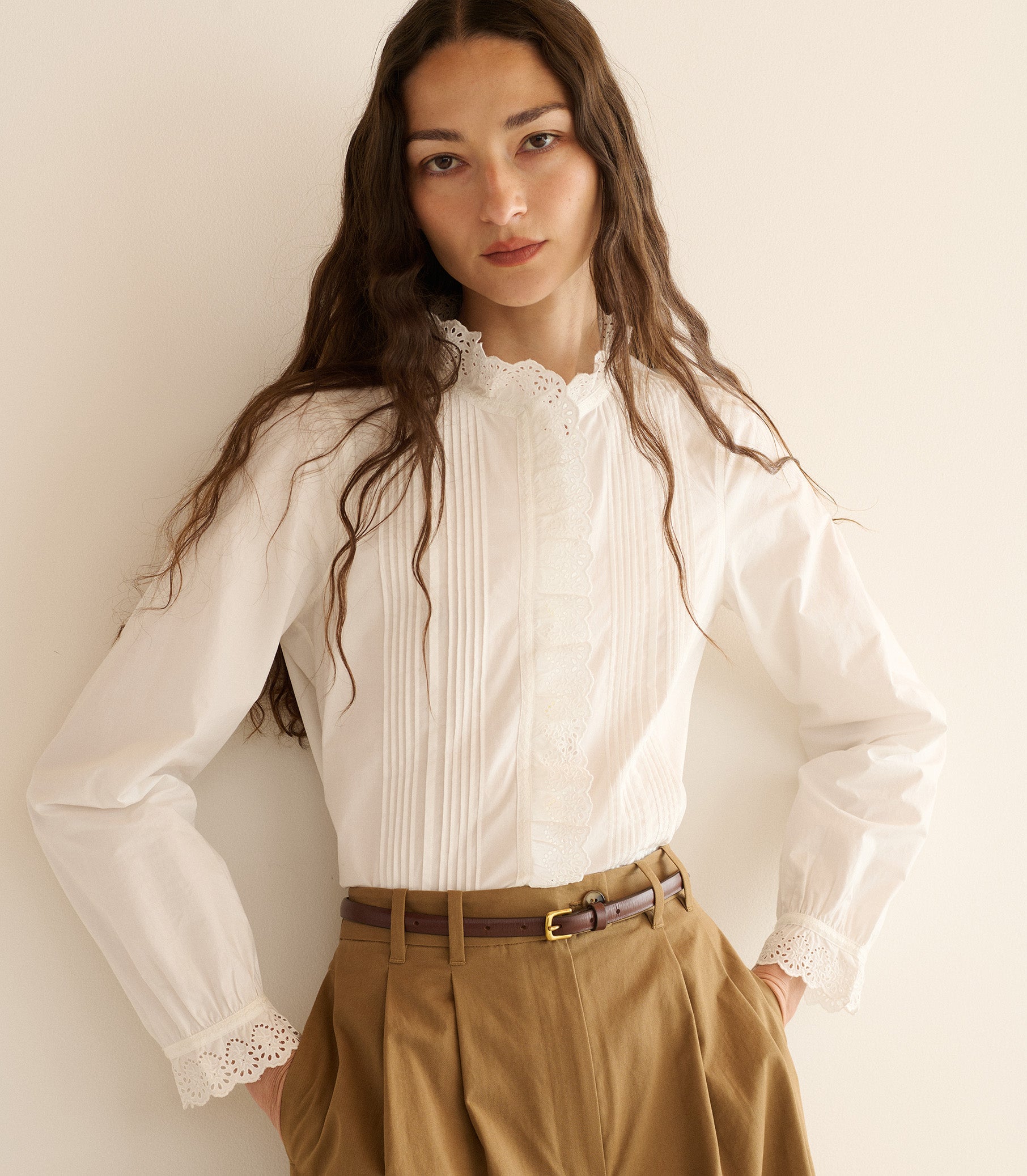 Woman wearing a white blouse with lace details and beige pants against a plain background