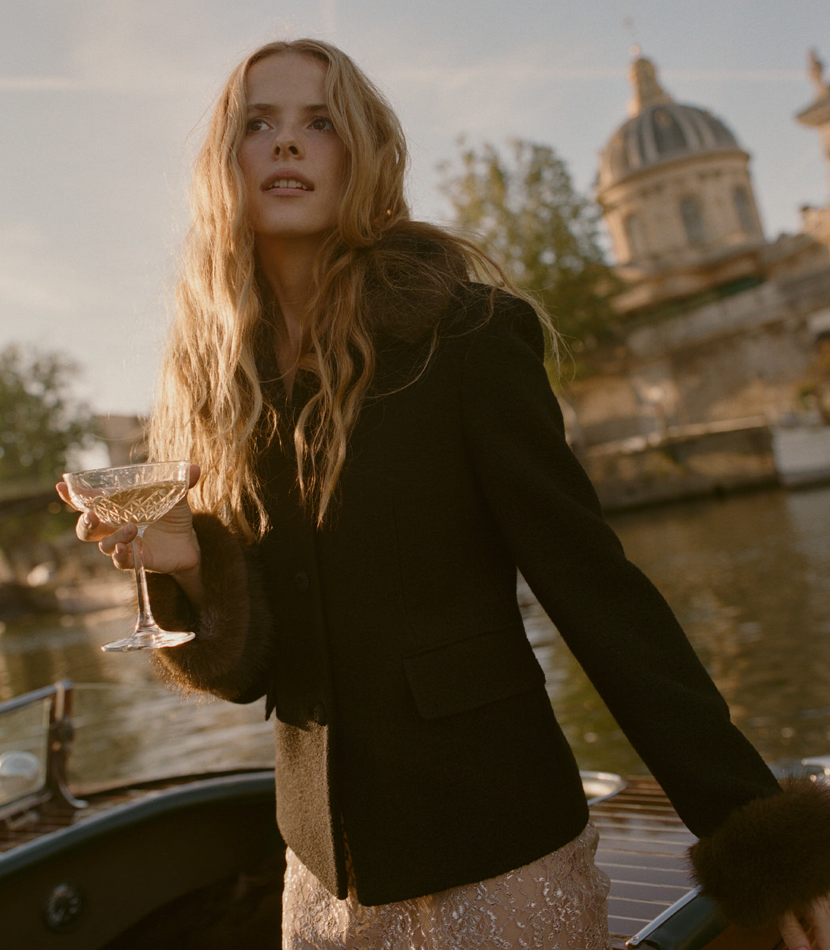 A woman with long blonde hair stands on a boat by a river, wearing a dark tailored jacket with dark fur trim and holding a coupe glass. A domed stone building is visible in the background. 2

