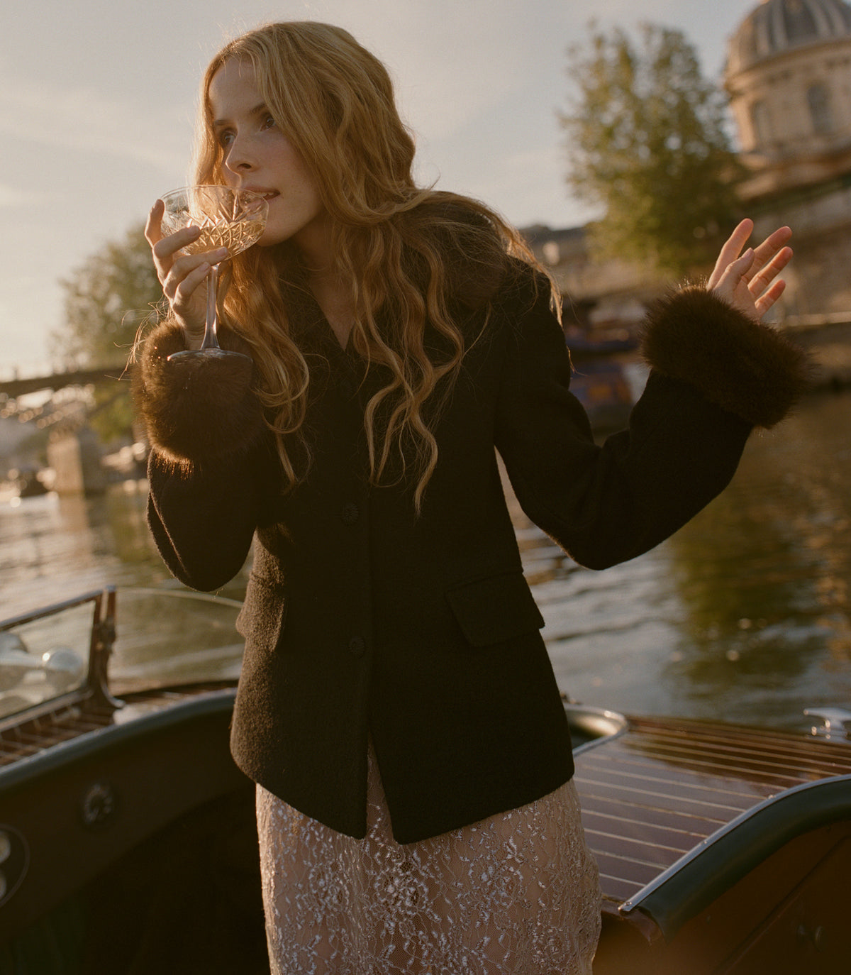 A woman stands on the bow of a boat at sunset, holding a wine glass. She is wearing a dark brown textured jacket with dark brown faux-fur trimmed cuffs and a champagne-colored sequined skirt.