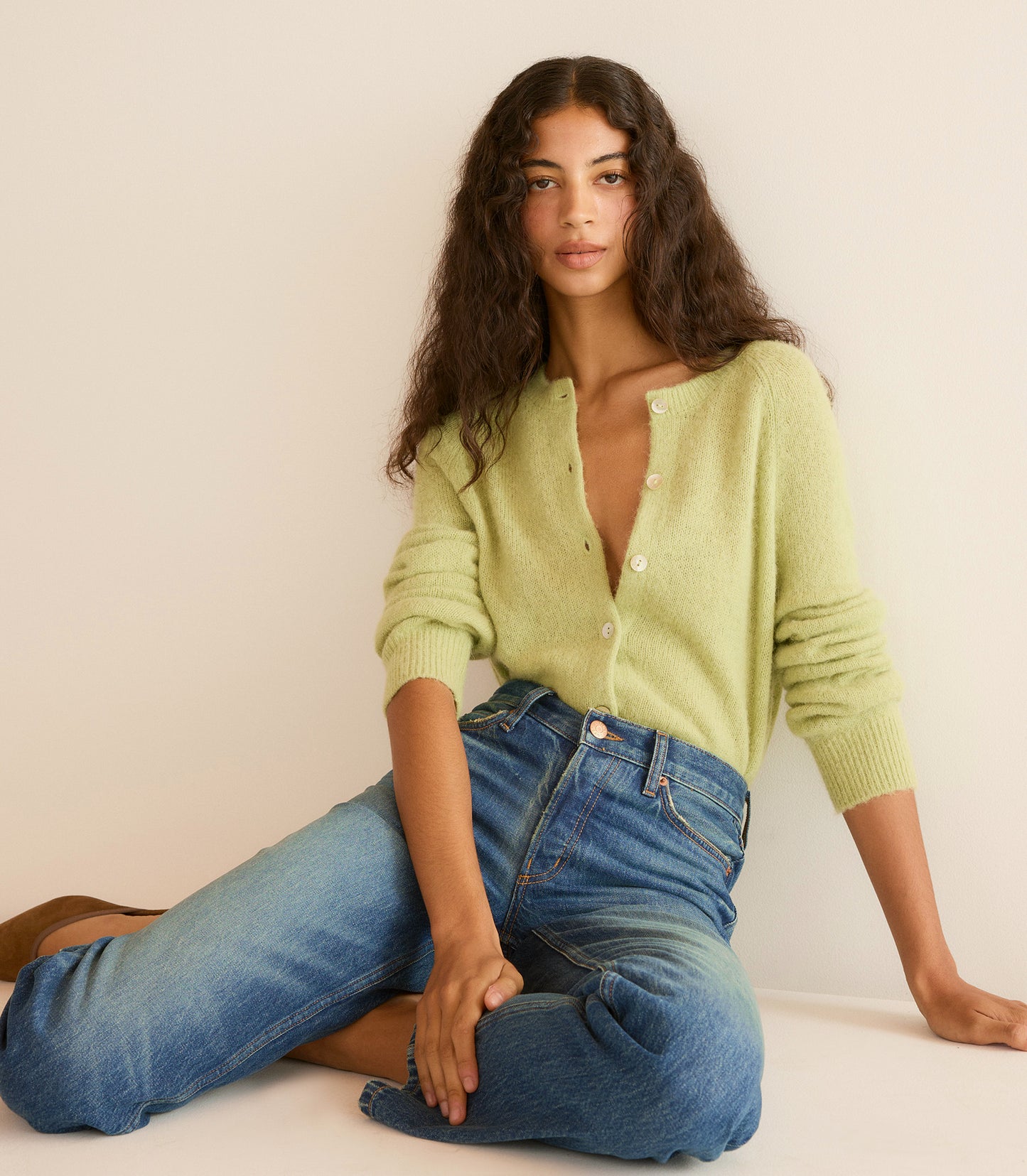 Woman wearing a light green cardigan and blue jeans sitting on the floor against a beige wall.